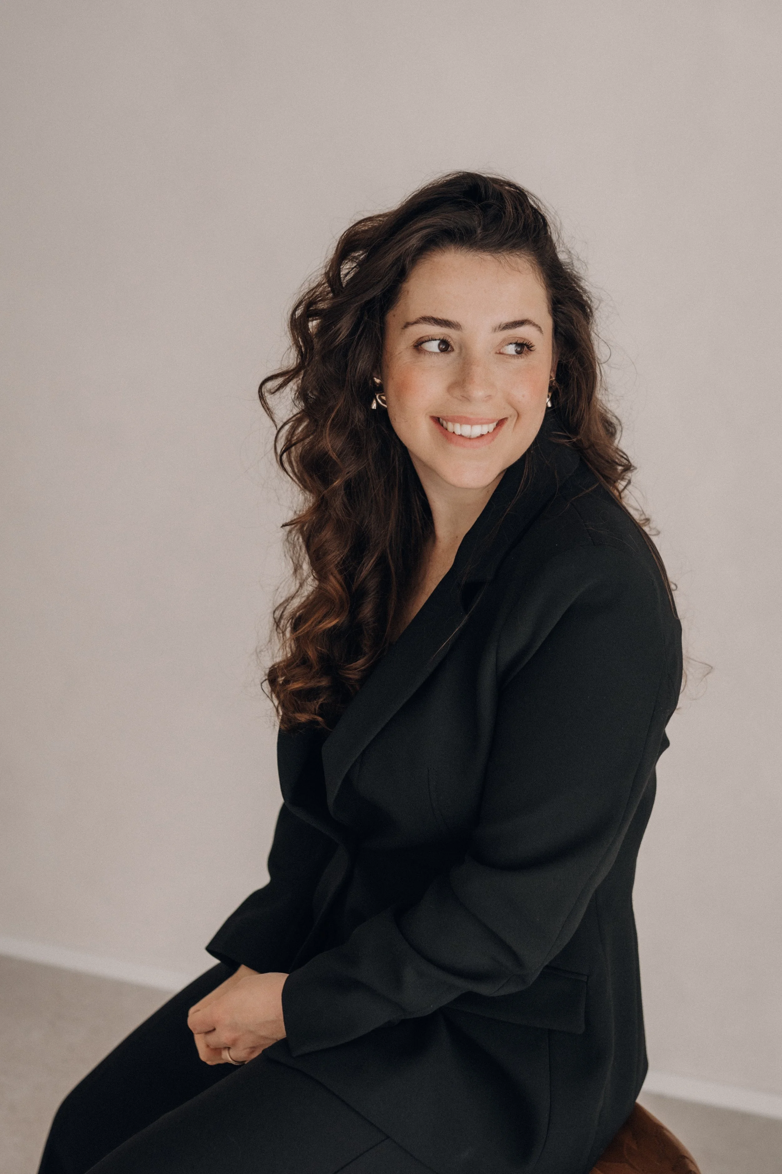 Jeune femme avec des cheveux bouclés bruns, portant un tailleur noir, assise, souriant et regardant vers sa gauche, dans un décor neutre.