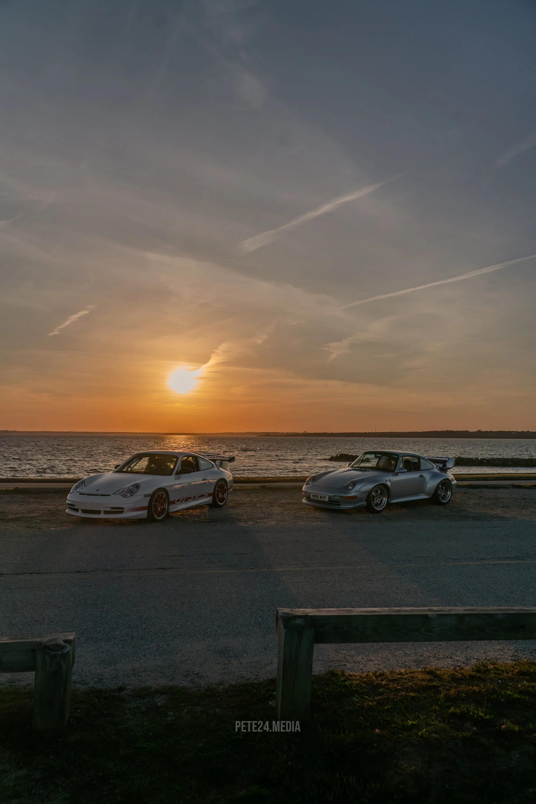 A silver Porsche 911 sports car parked near a body of water during sunset, with a sign indicating parking reservation nearby.