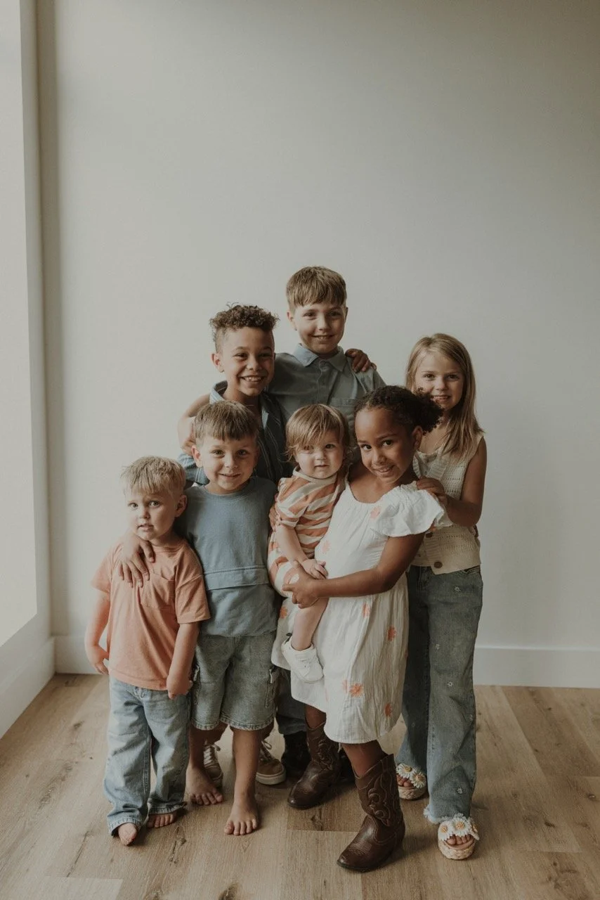 Group of seven children of diverse ethnicities standing together indoors, smiling, with some hugging each other.