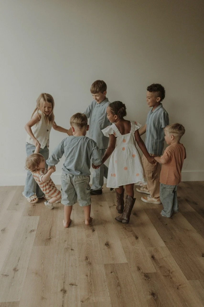 A diverse group of seven children holding hands and forming a circle, playing together indoors on a wooden floor against a plain white wall.