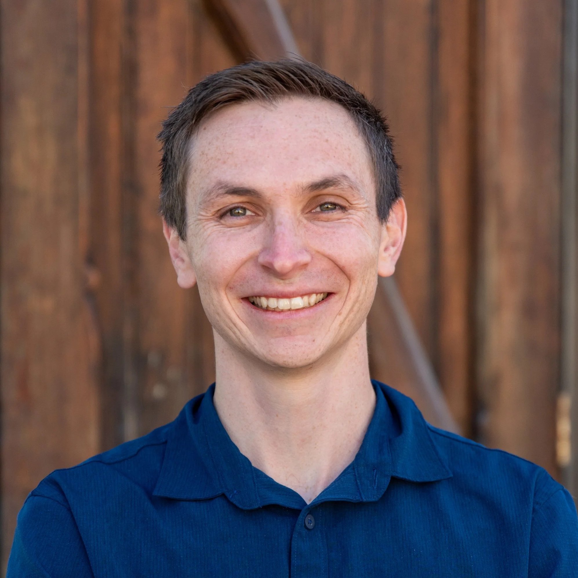 A man with curly dark hair, glasses, and a beard, wearing a dark green sweater, looking thoughtfully to the side, with a blurred plant in the background.