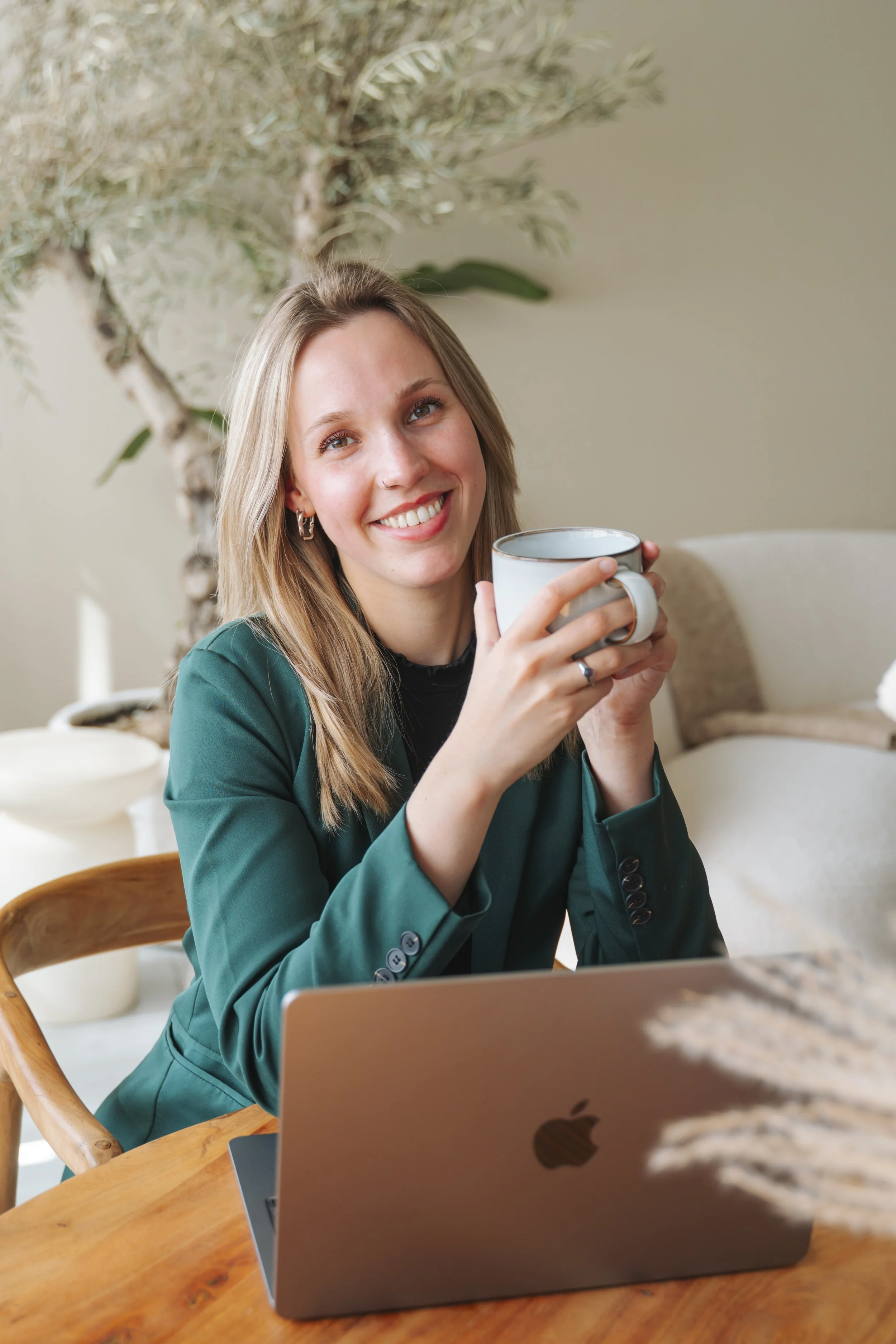 Vrouw met blonde haar die hét voor een kop koffie. Ze zit aan een houten tafel met een laptop en een groene jas. Achter haar is een plant en een witte bank.