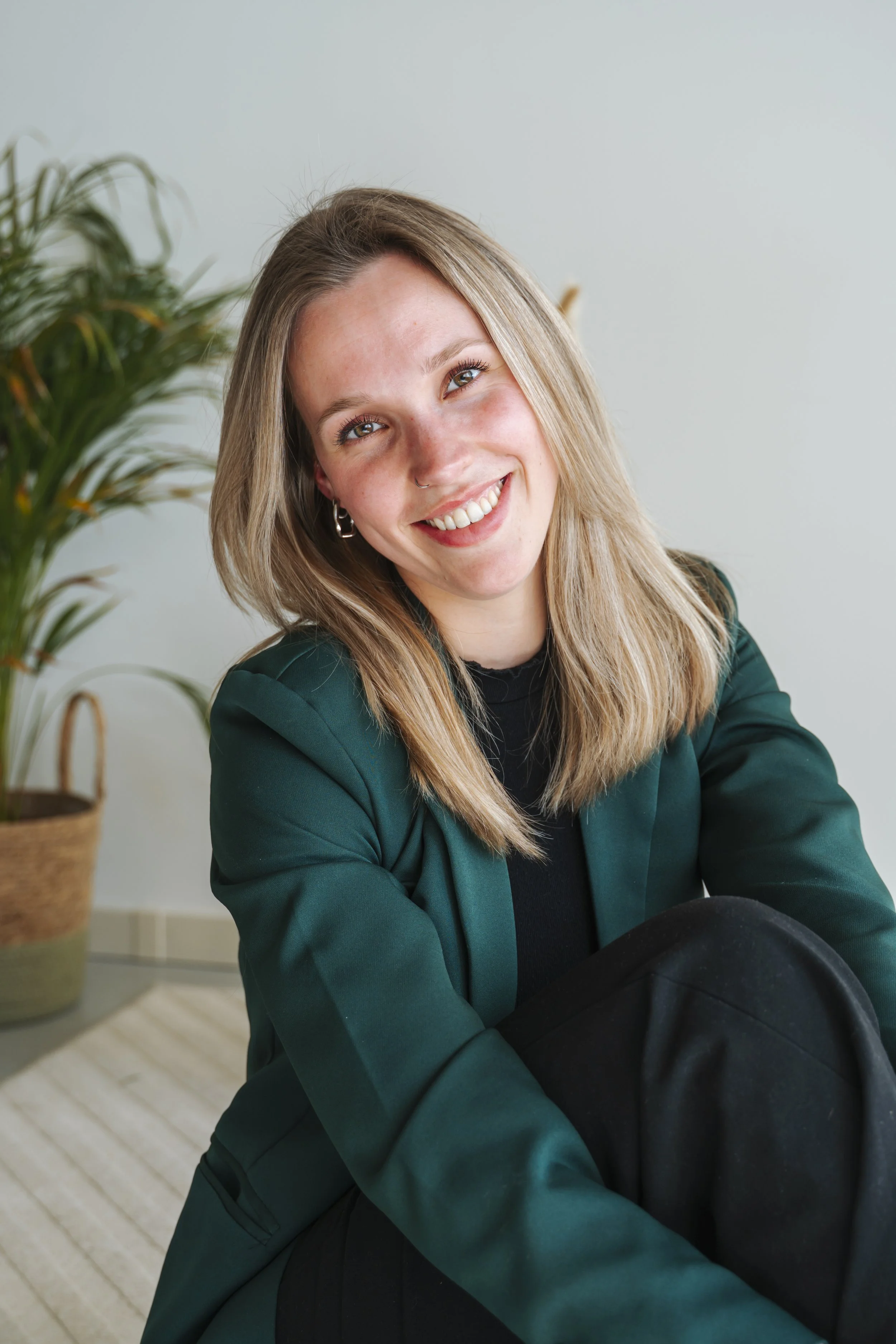 Vrouw met lang blond haar, glimlachend, zittend in een kamer met een plant op de achtergrond.