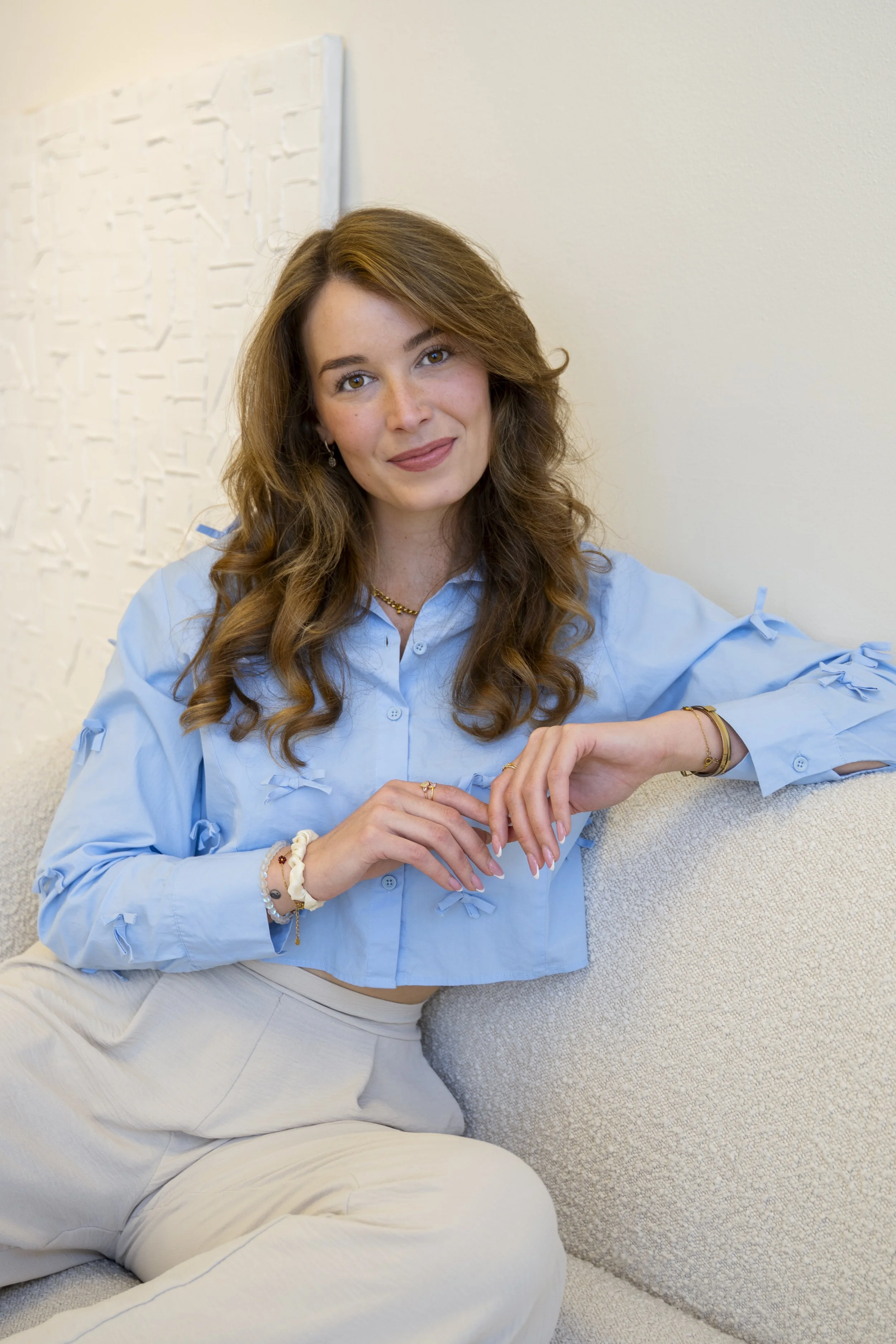 Een jonge vrouw met bruin, golvend haar in een blauwe blouse en lichte broek zit op een bank en lacht naar de camera.