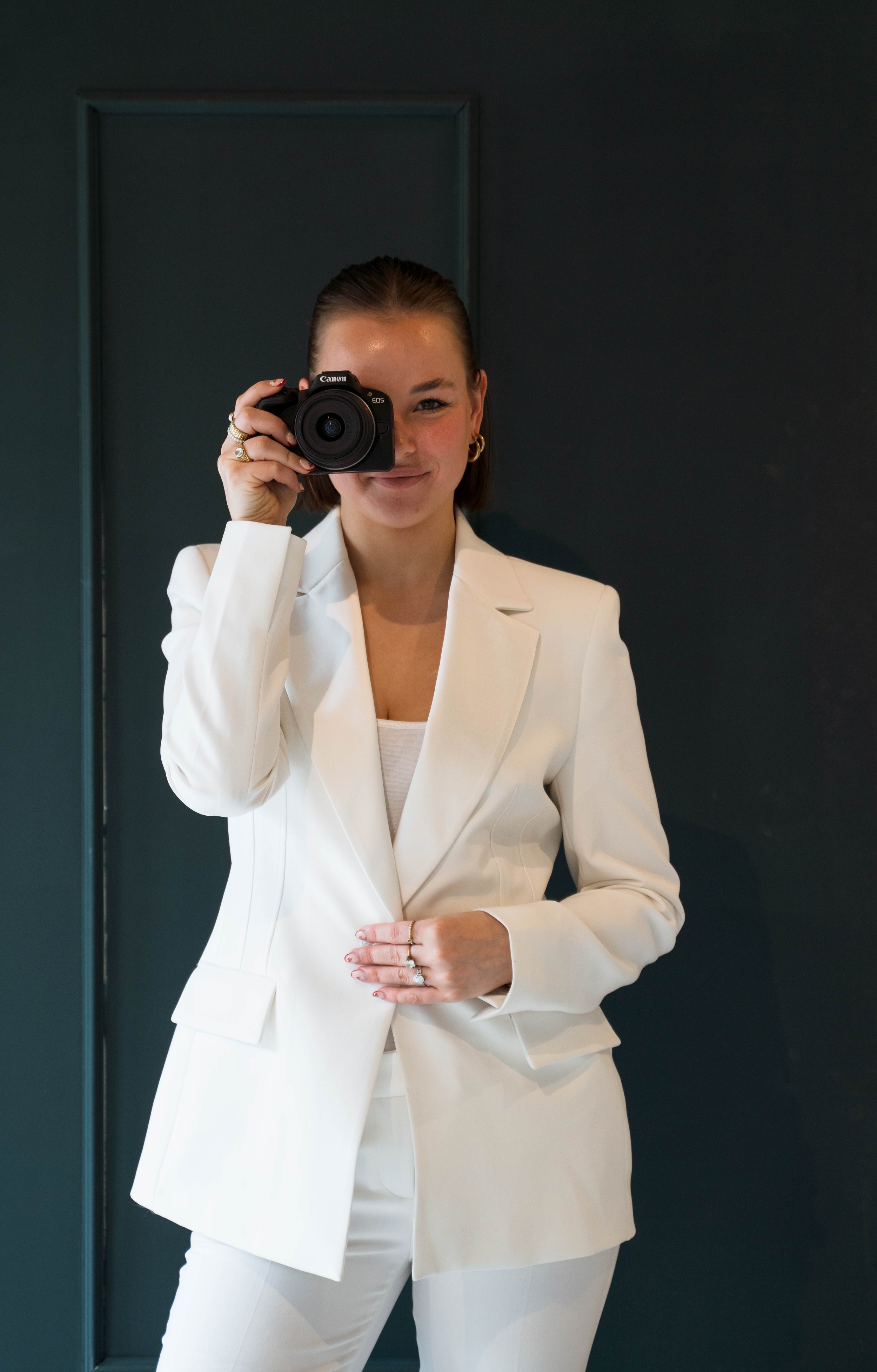 Vrouw in witte pak die een camera vasthoudt en door een spiegel zichzelf fotografeert