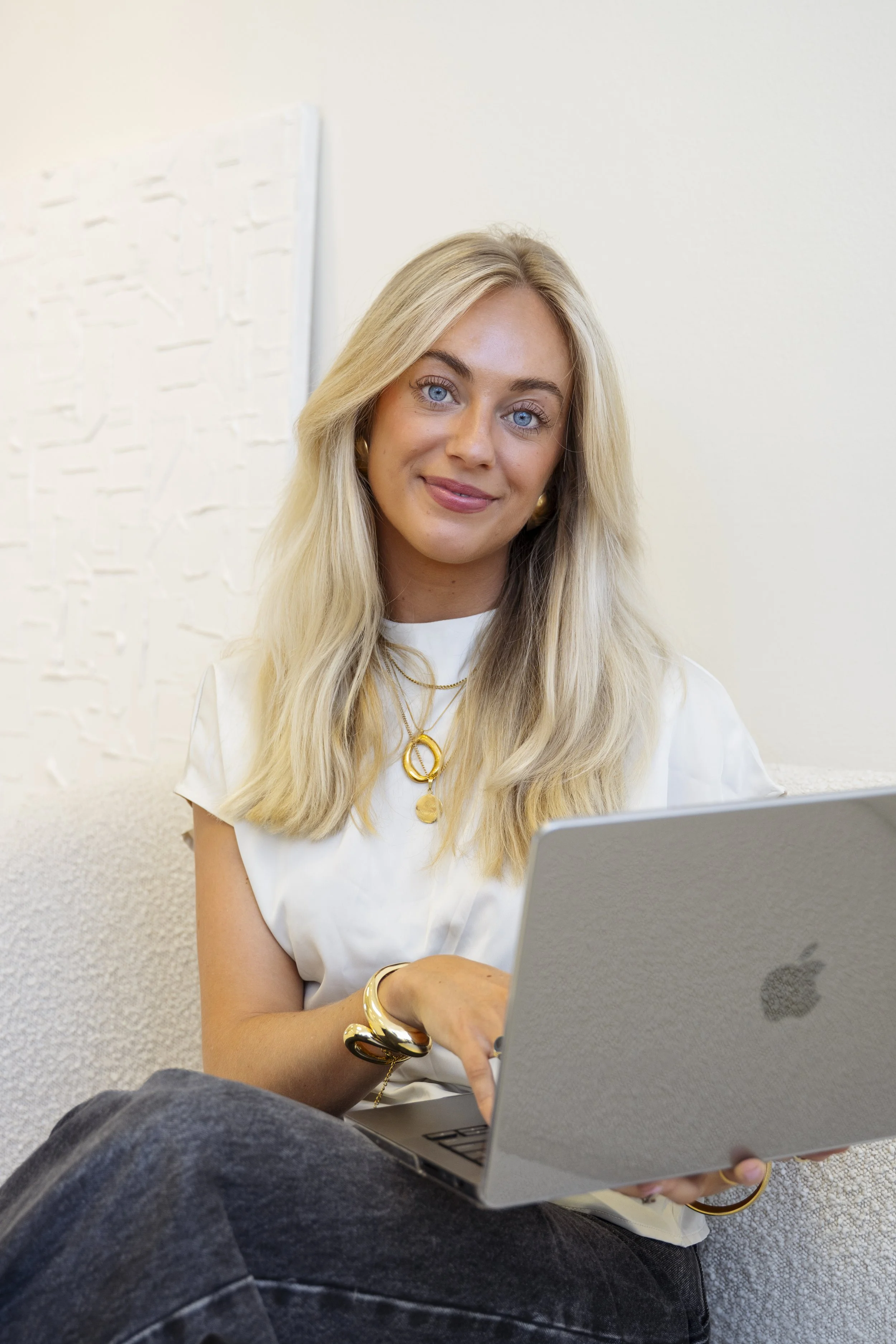 Een lachende blonde vrouw met blauwe ogen zit op een bank, gebruikt een zilveren MacBook, draagt een witte top, gouden sieraden en heeft lang, golvend haar.