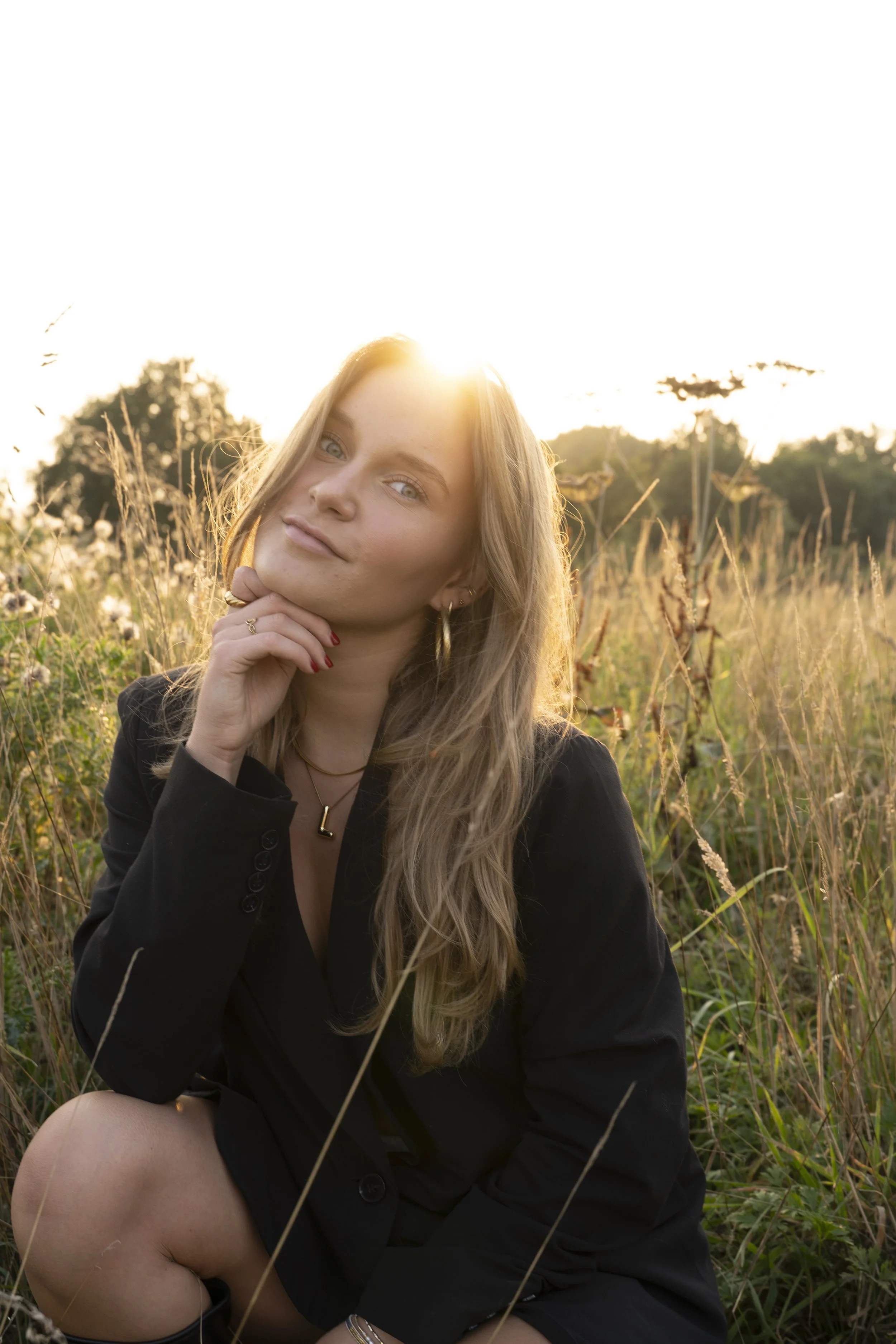Portret van een jonge vrouw met lang blond haar, zittend in een veld met droog gras tijdens zonsondergang, met de zon achter haar hoofd.