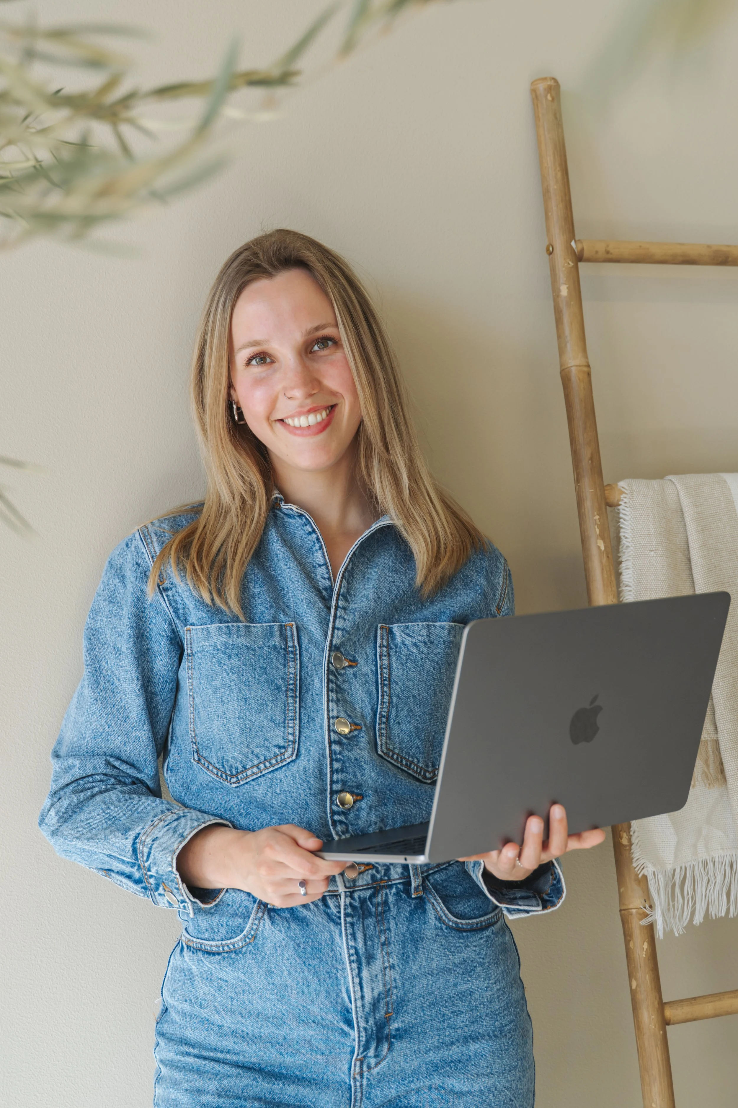 Vrouw met lange blonde haar in een denim jas en rok, die een MacBook vasthoudt, glimlachend voor een neutrale muur met een houten ladder en handdoek op de achtergrond.