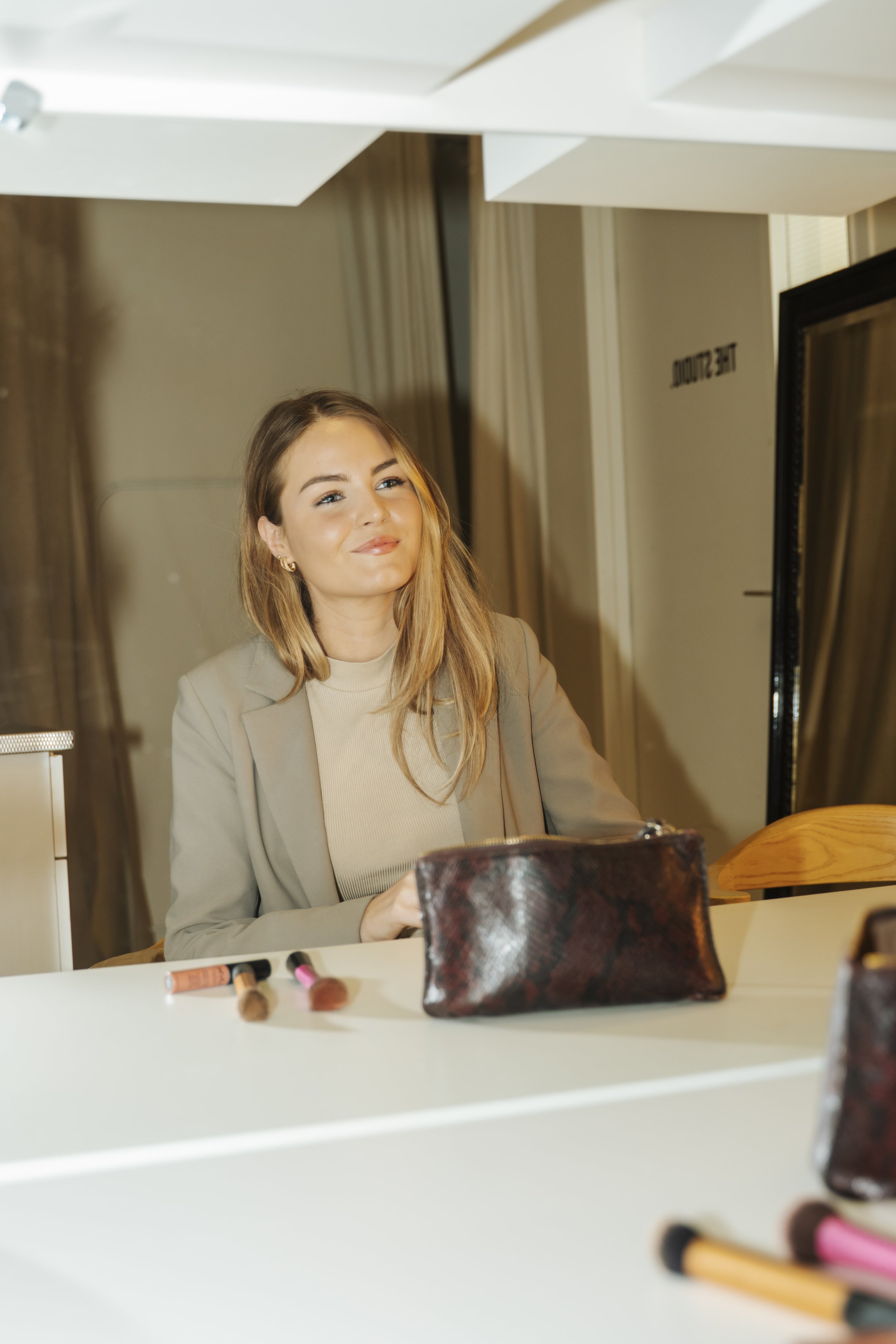 Een vrouw met lang blond haar zit aan een tafel met make-up producten, glimlachend.