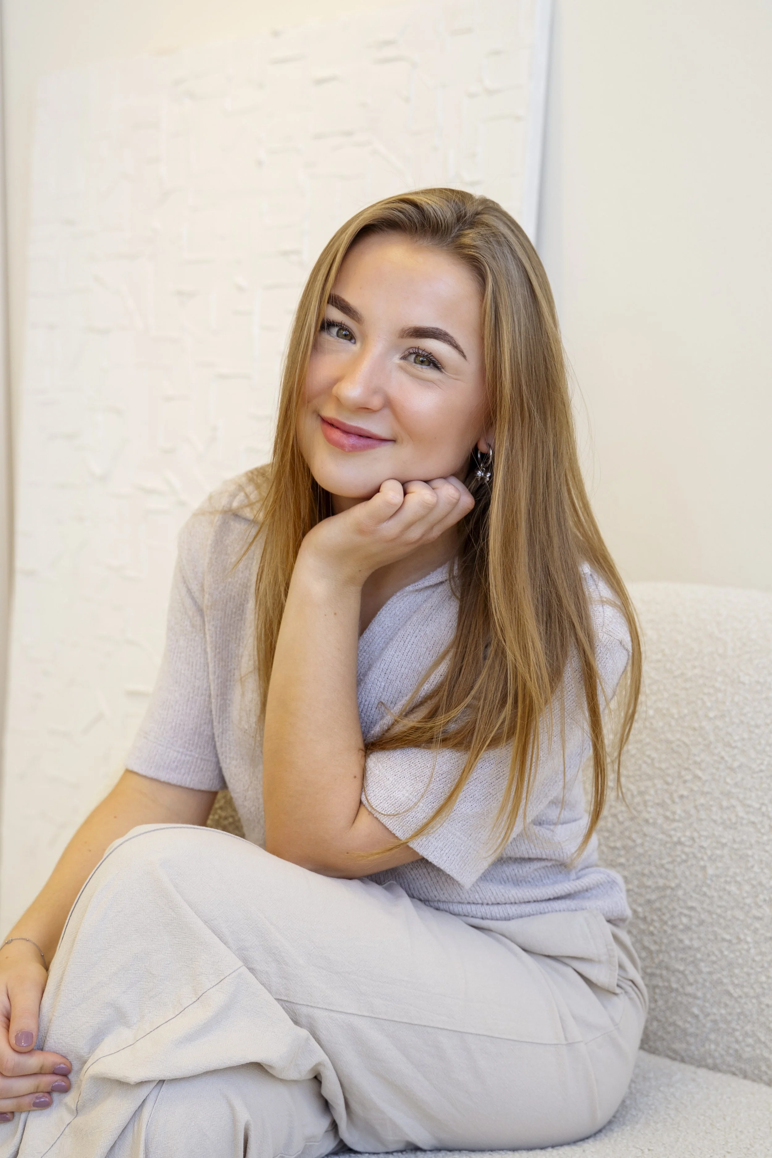 Een jonge vrouw met lang, kastanjebruin haar glimlacht, rust haar kin op haar hand, zittend op een lichte bank in een lichte kamer met witte muur.
