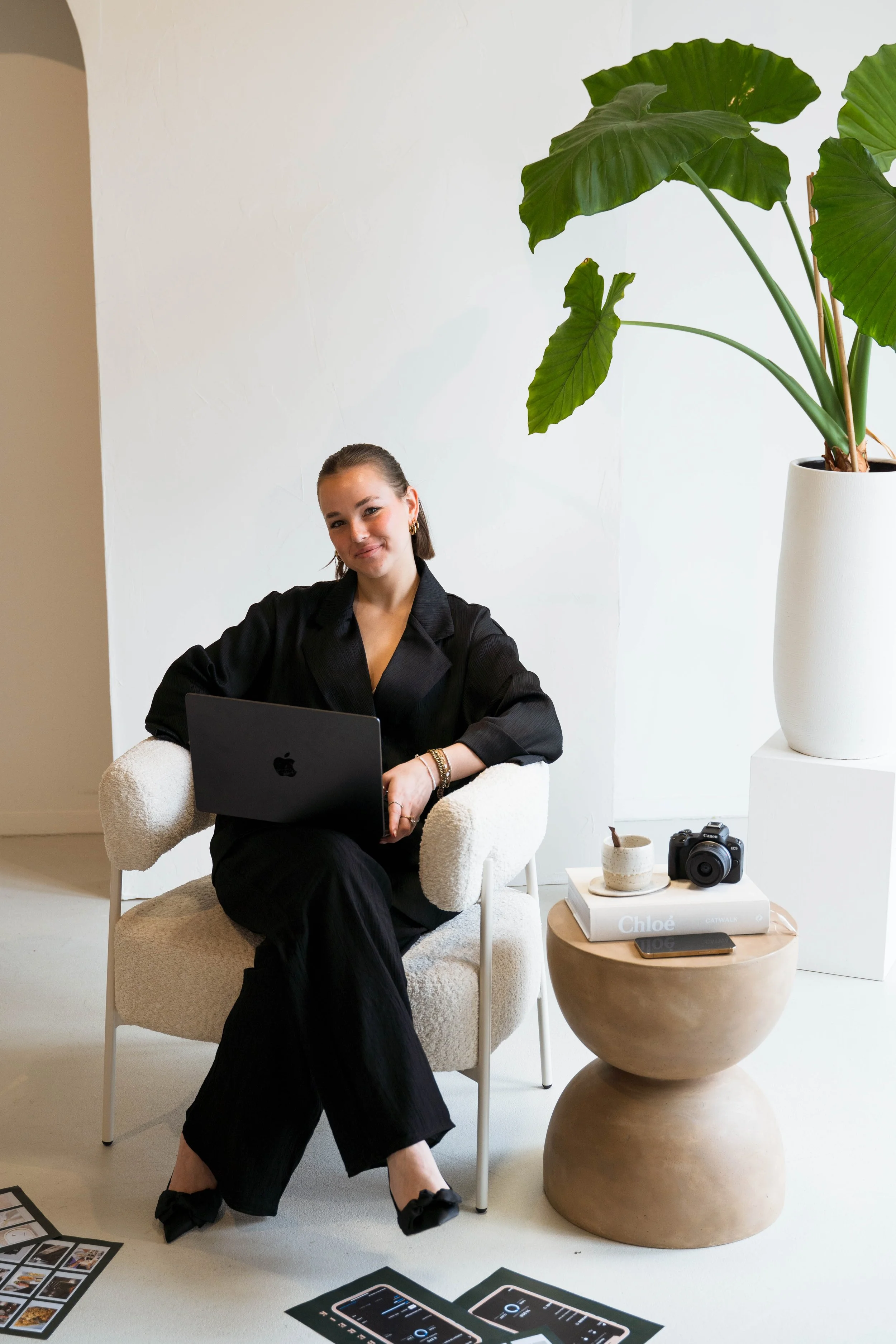 Vrouw zit in een witte kamer met een laptop op haar schoot, een grote groene plant op een wit voetstuk, een ronde bijzettafel met boeken, een camera, een mok en een telefoon, en er liggen foto's op de vloer.