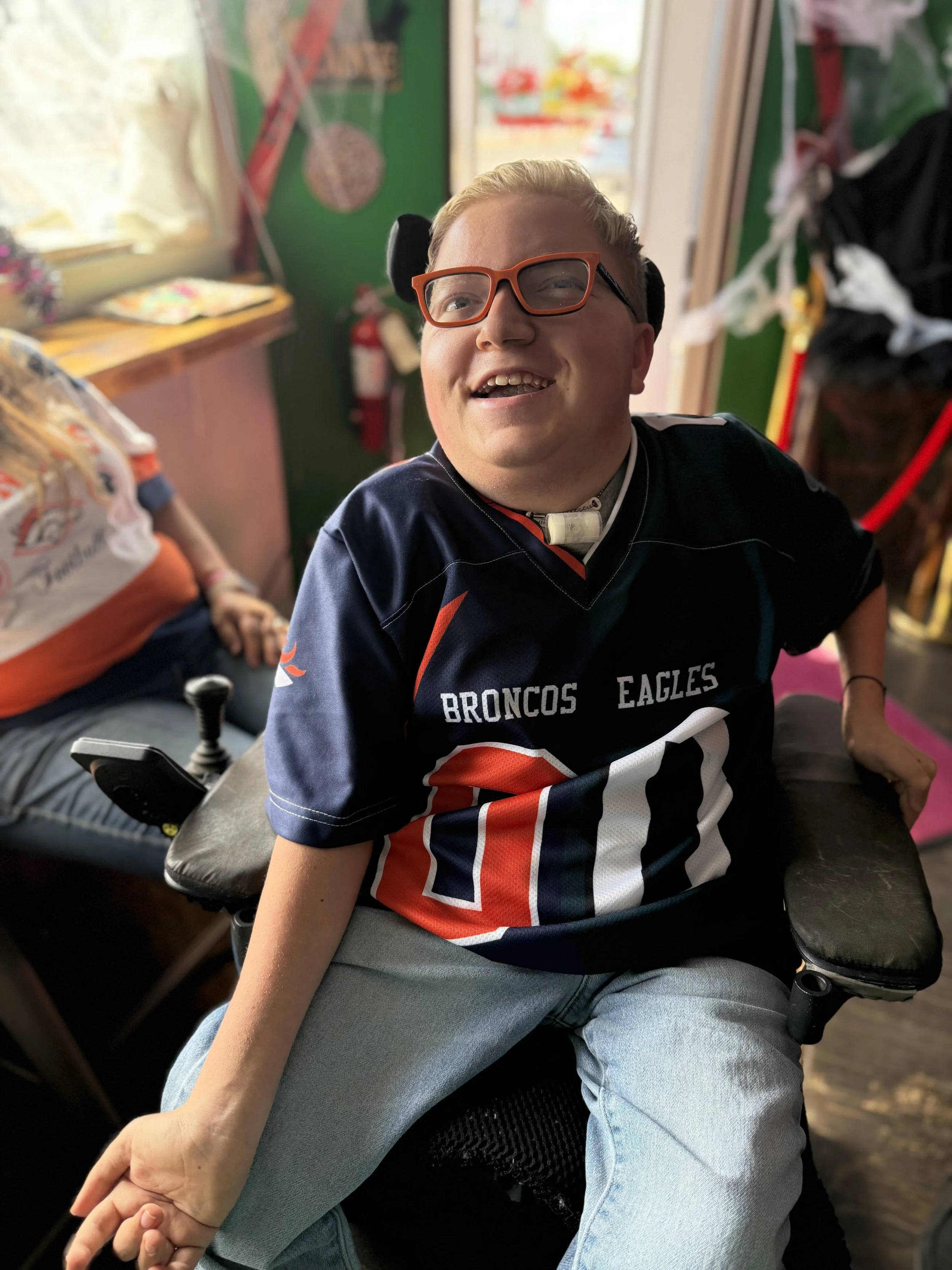 A young man with glasses smiling while sitting in a wheelchair indoors. He is wearing a Denver Broncos football jersey and light blue jeans.