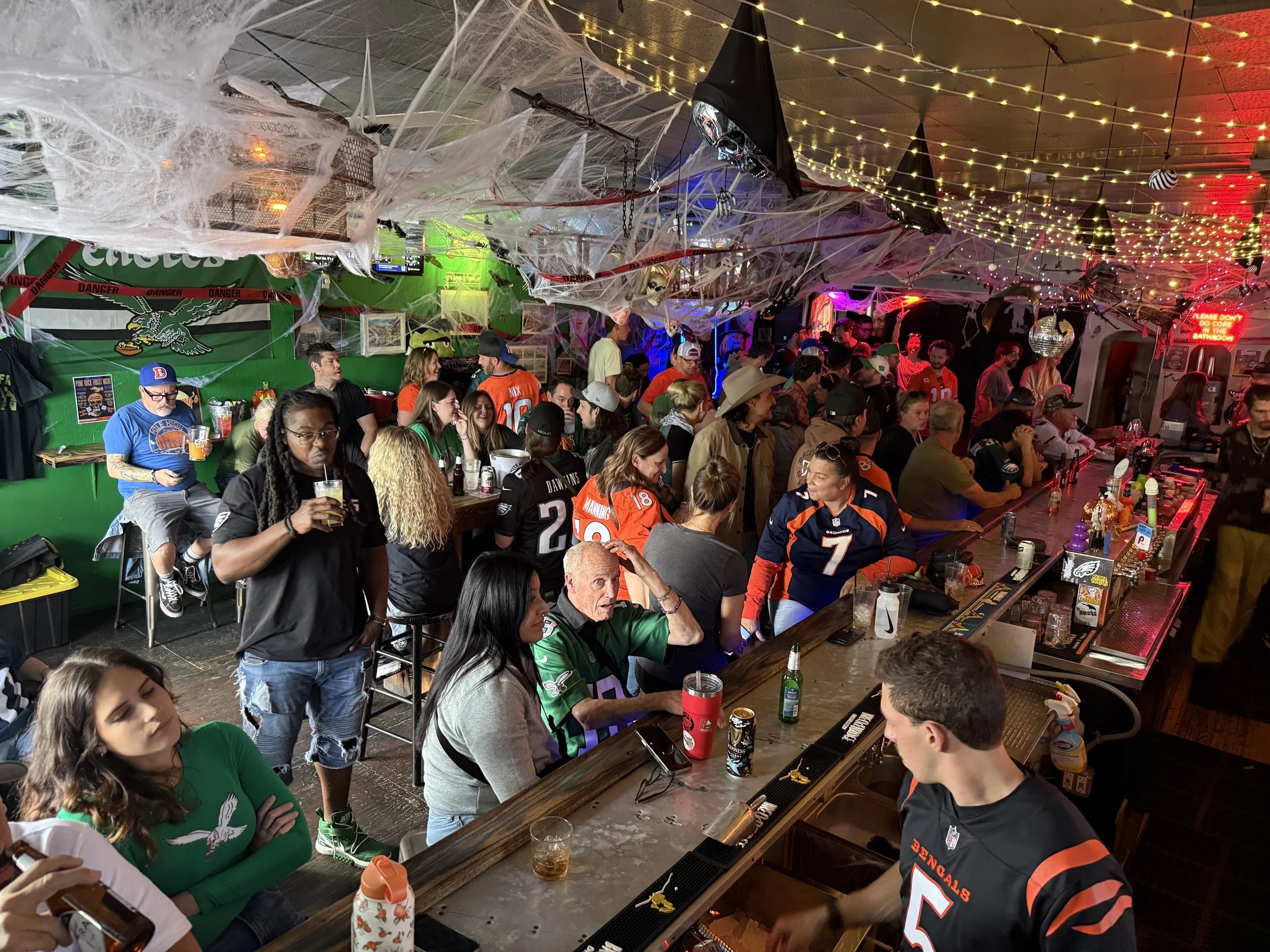 A crowded bar decorated with Halloween and sports themes, featuring spider webs, hanging bats, string lights, and jerseys of various football teams, with people socializing and drinking.