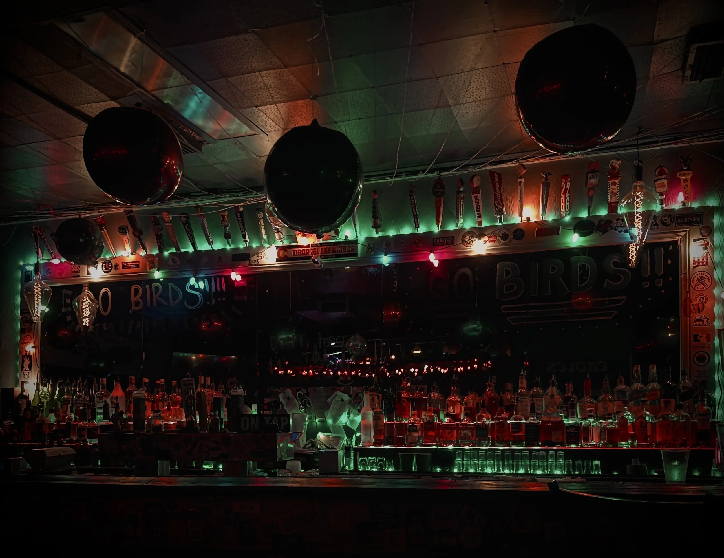 A bar decorated for a celebration with green and red lights, black balloons, and various bottles of alcohol on the counter. There are signs and decorations, including a 'GO BIRDS' sign and many sports-themed stickers on the wall behind the bar.