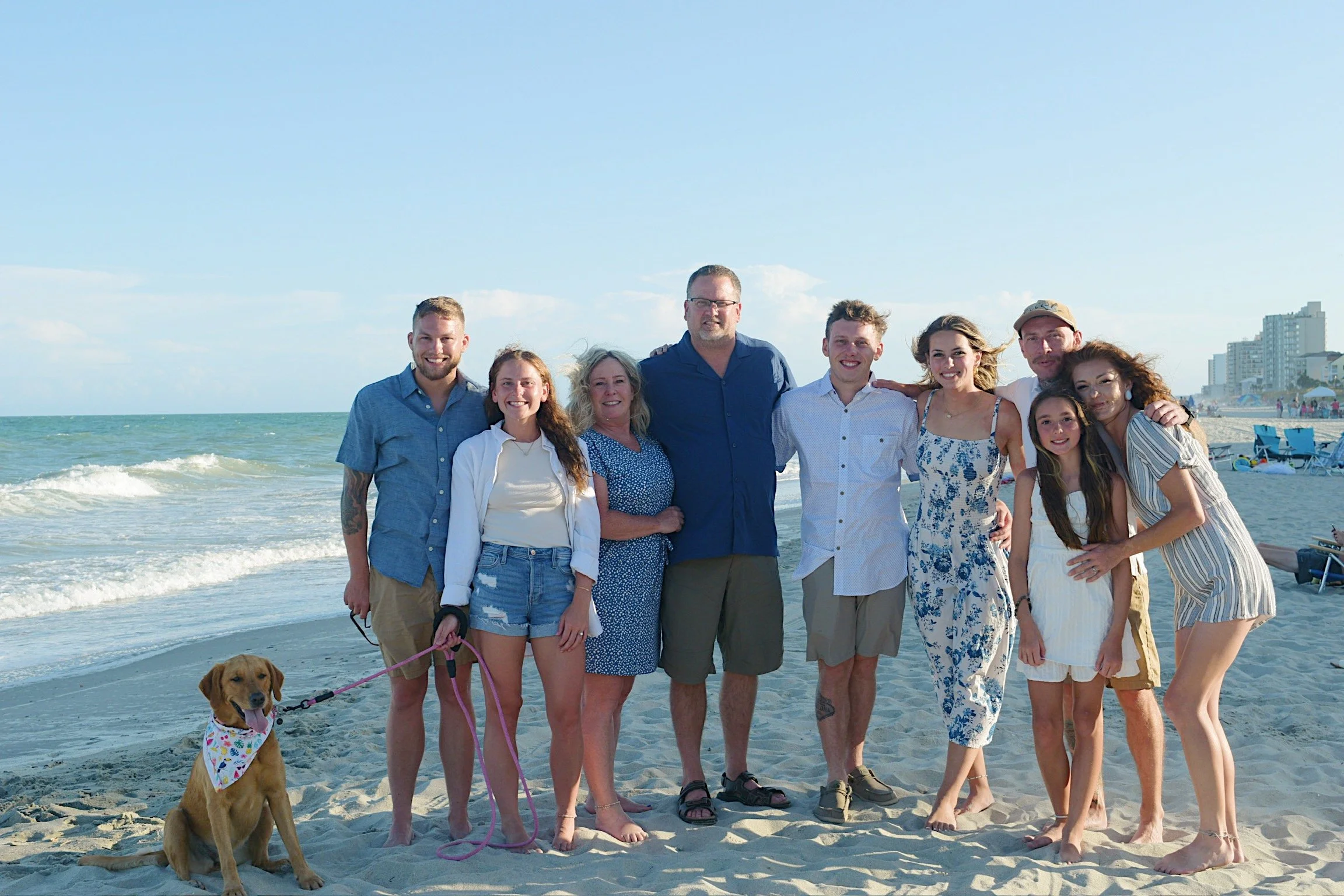 A group of ten people, including children and adults, and a dog, standing on a sandy beach near the ocean with waves, with beach chairs and buildings in the background, under a partly cloudy sky.