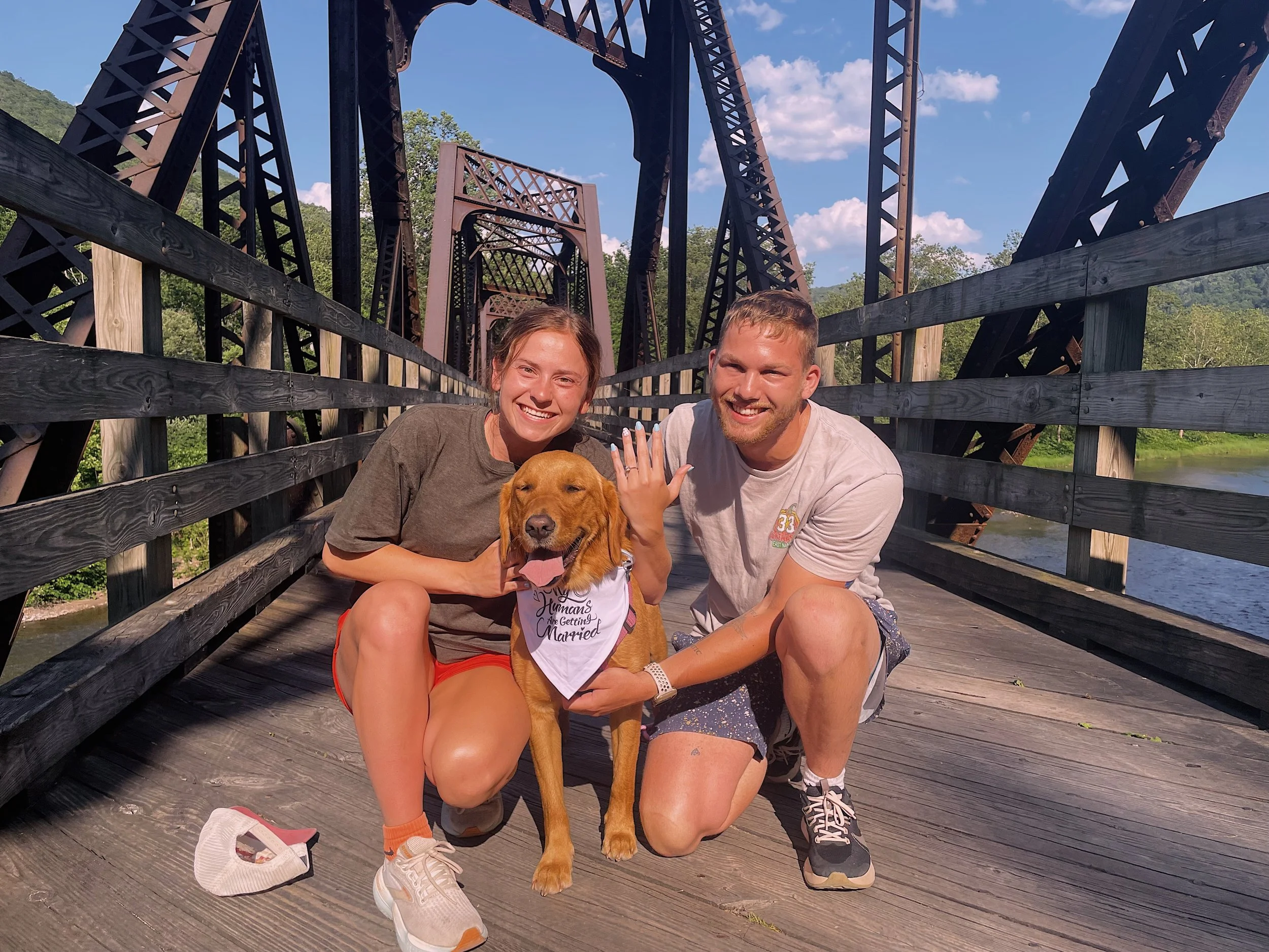 A man and woman kneeling on a wooden bridge with a dog between them, during daytime. The woman is showing her left hand with an engagement ring, and the dog is wearing a bandana that says 'Humans Are Getting Married.' The bridge has black metal arches and wooden railings, with greenery and a river visible in the background.