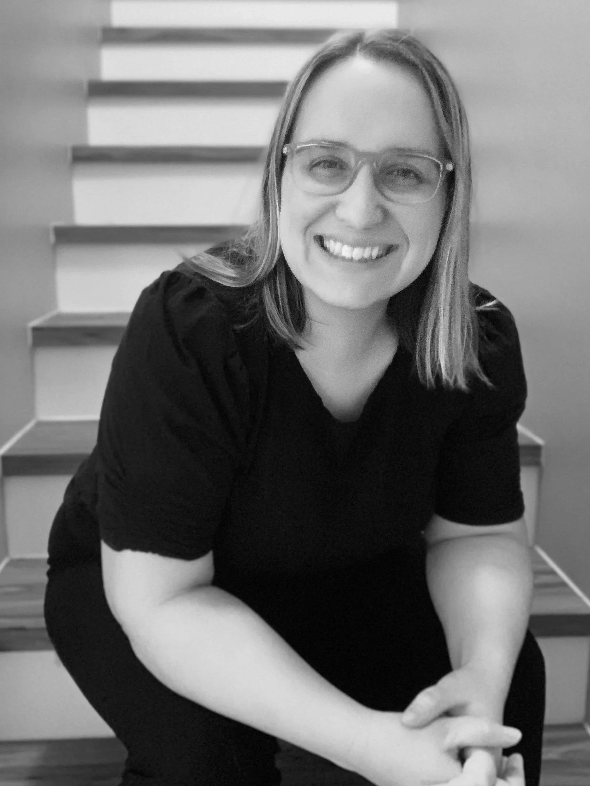 A smiling woman with glasses and shoulder-length hair, sitting on stairs with a wooden floor in the background.