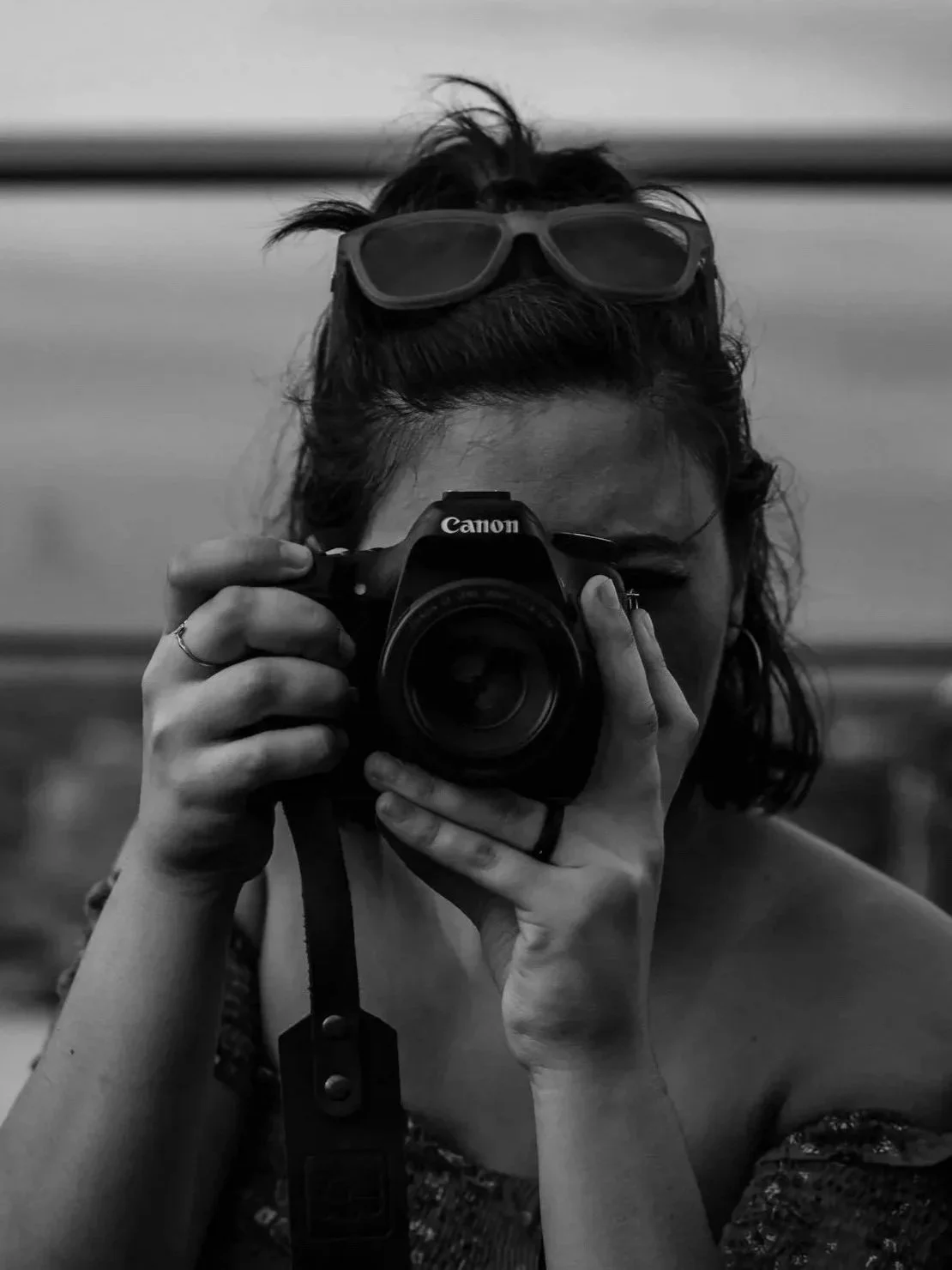 A person with sunglasses on their head holding a Canon camera to their face, taking a photo in black and white.