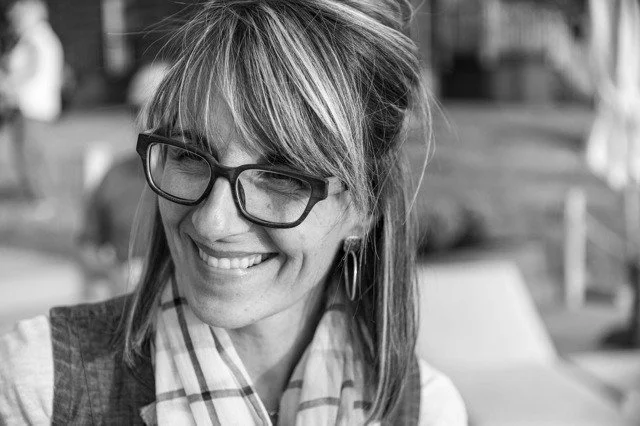 A woman with glasses smiling, wearing a scarf and earrings, in an outdoor setting.