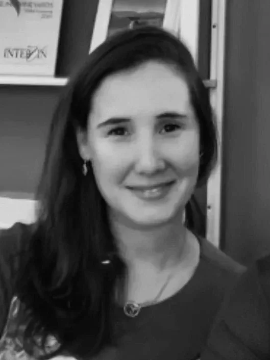 Black and white photo of a woman with long dark hair, smiling, wearing a necklace and earrings, in an indoor setting with shelves and picture frames in the background.