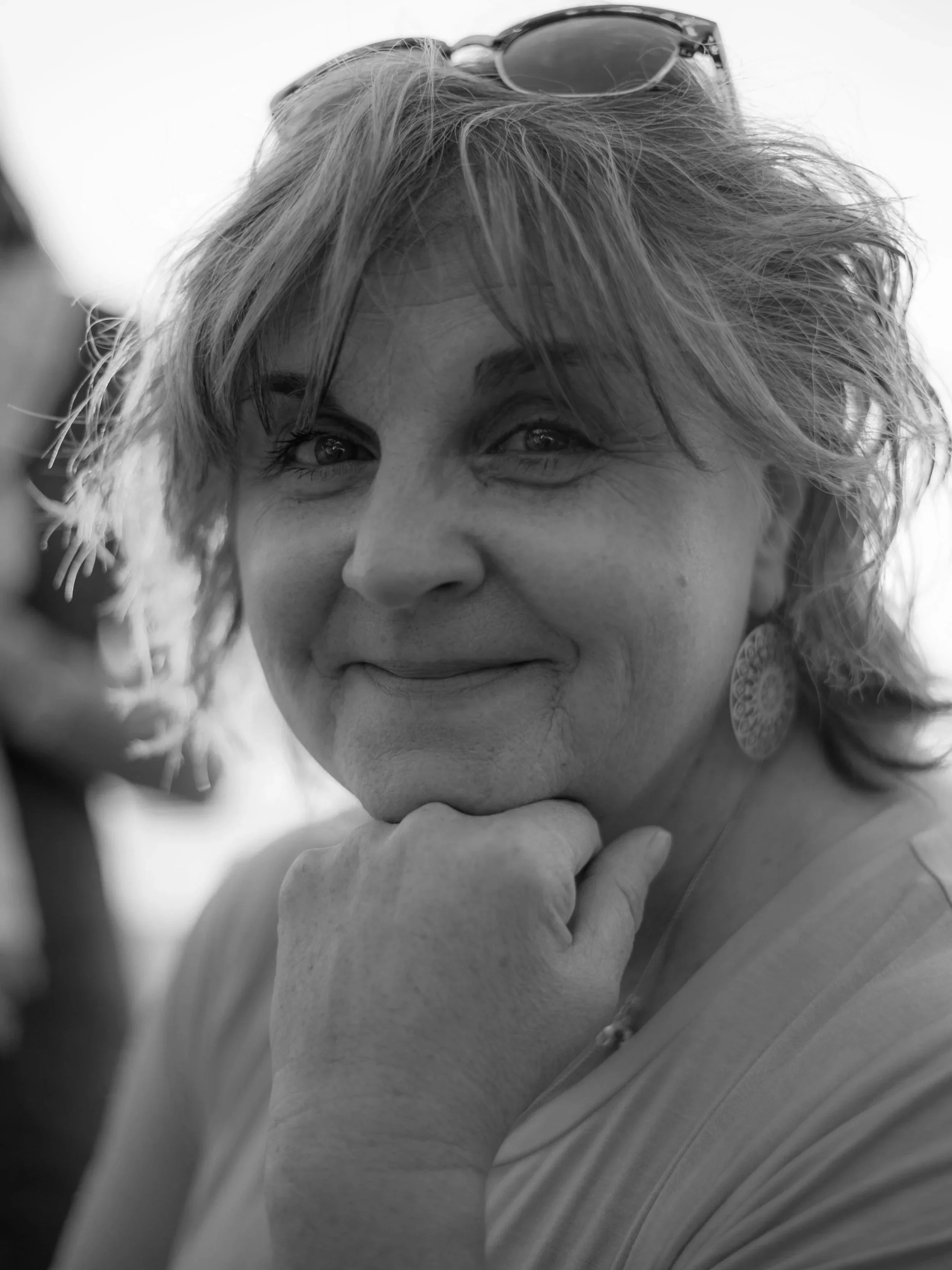 Black and white photo of a woman with short, curly hair, wearing sunglasses on her head, hoop earrings, and a light-colored top, resting her chin on her hand with a slight smile.