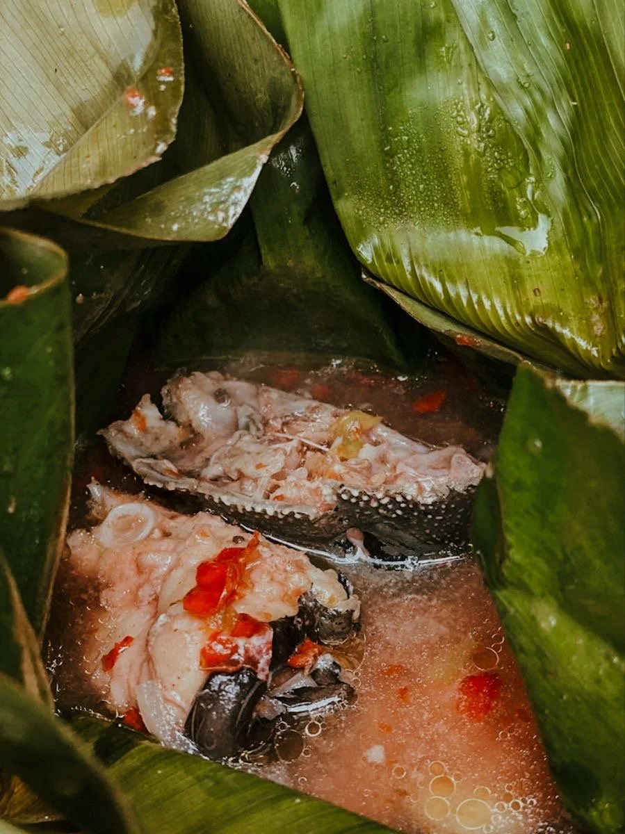 Une préparation culinaire dans des feuilles de bananier, contenant du poisson cuit dans une sauce tomate.