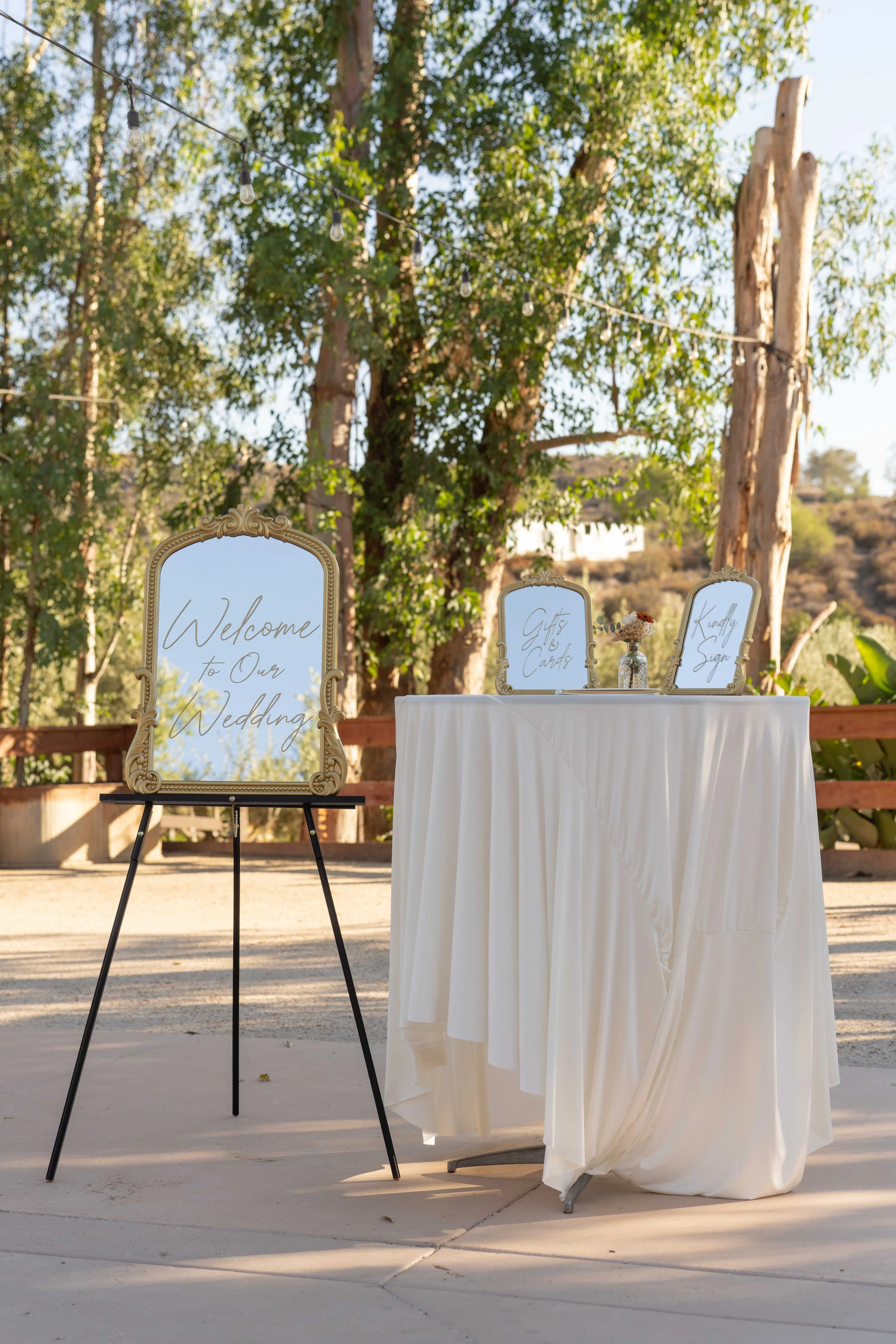 Wedding reception table with signage and a welcome sign, outdoors with trees and string lights.