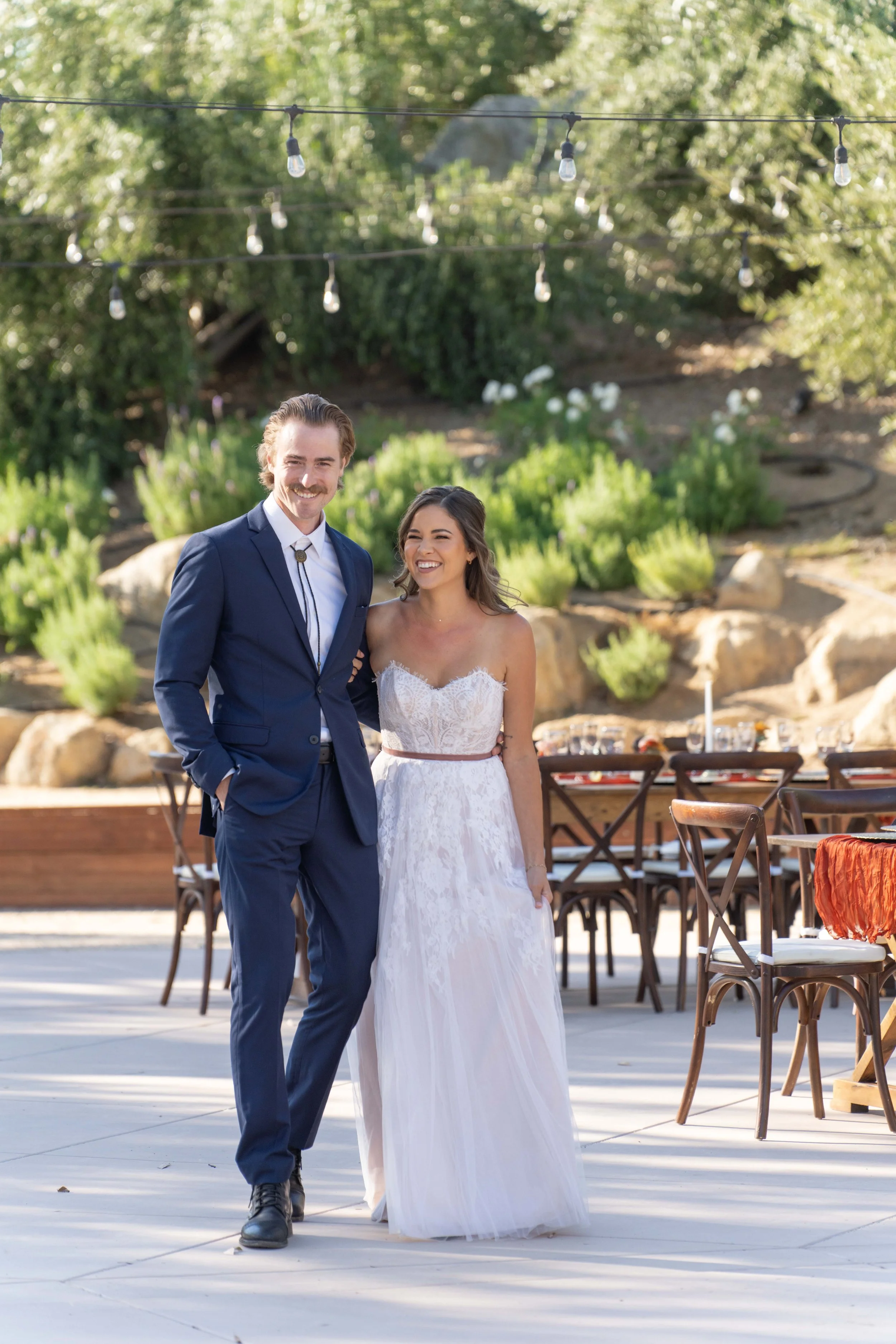 A happy couple at a wedding reception outdoors, with string lights overhead and a garden background, dressed in wedding attire.