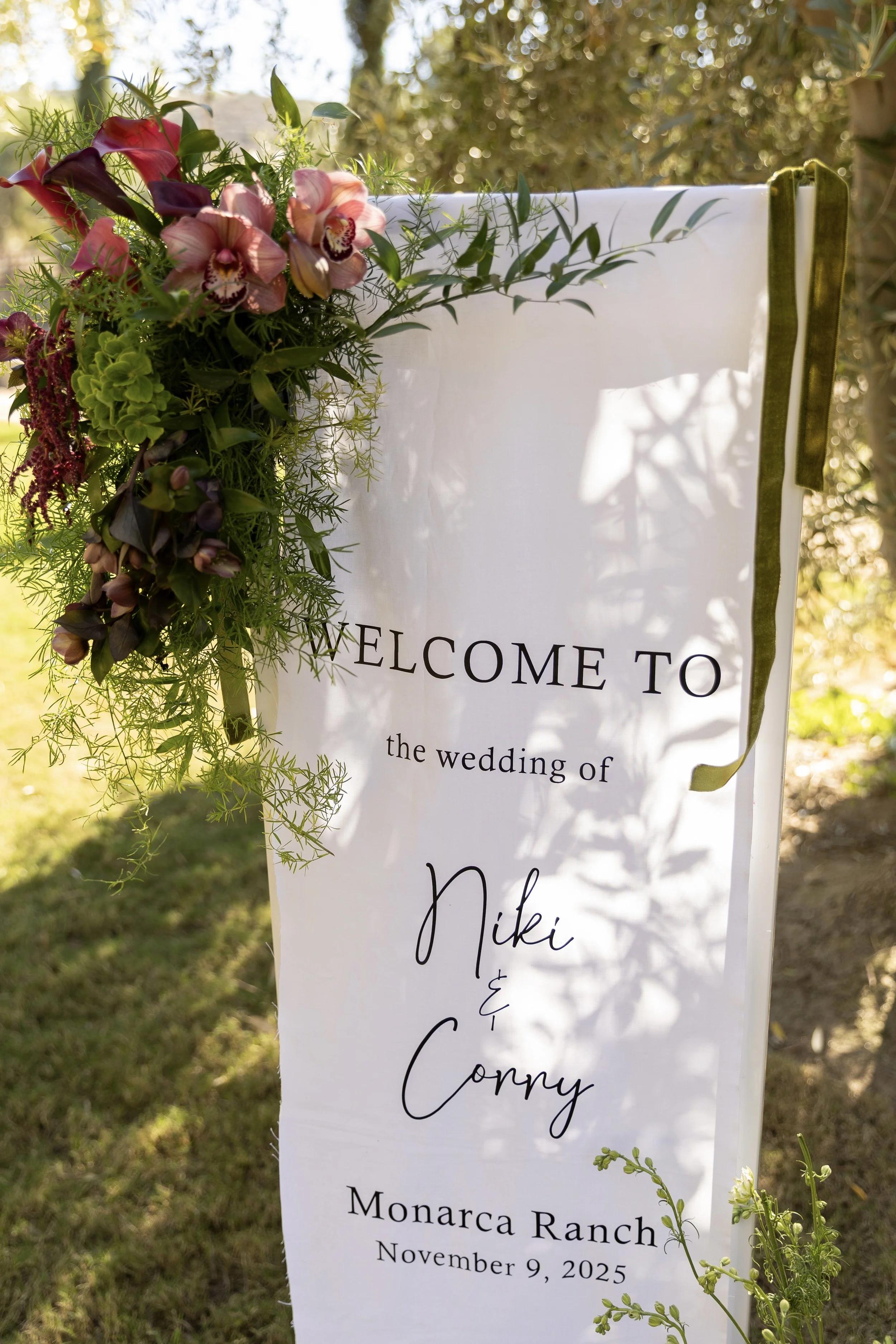 Wedding welcome sign decorated with green foliage and purple flowers, displaying the names Nikki and Coppy at Monarca Ranch on November 9, 2025.