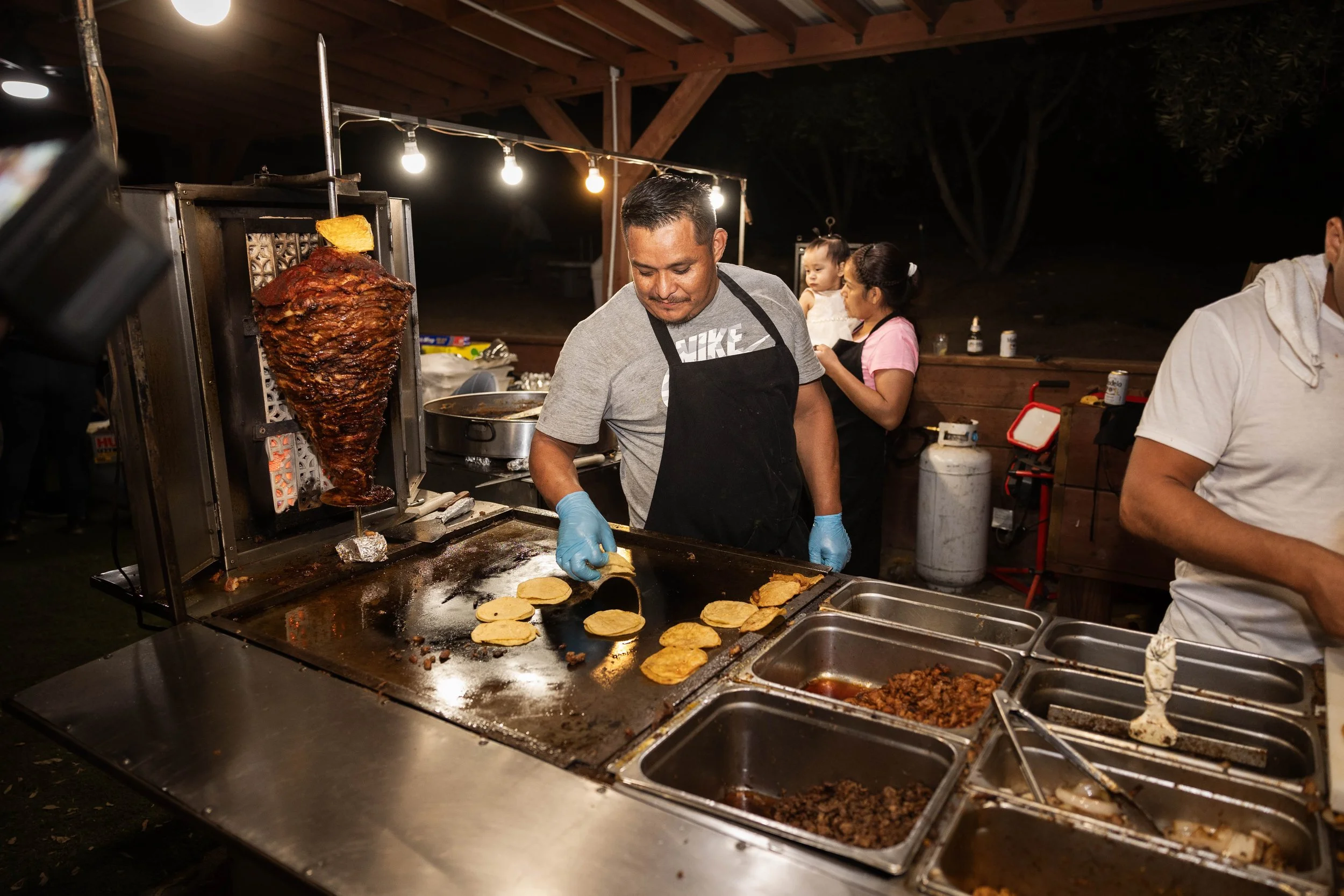 A man cooking tacos on a flat-top grill with a large vertical spit of meat behind him, and a few people in the background at an outdoor night event.