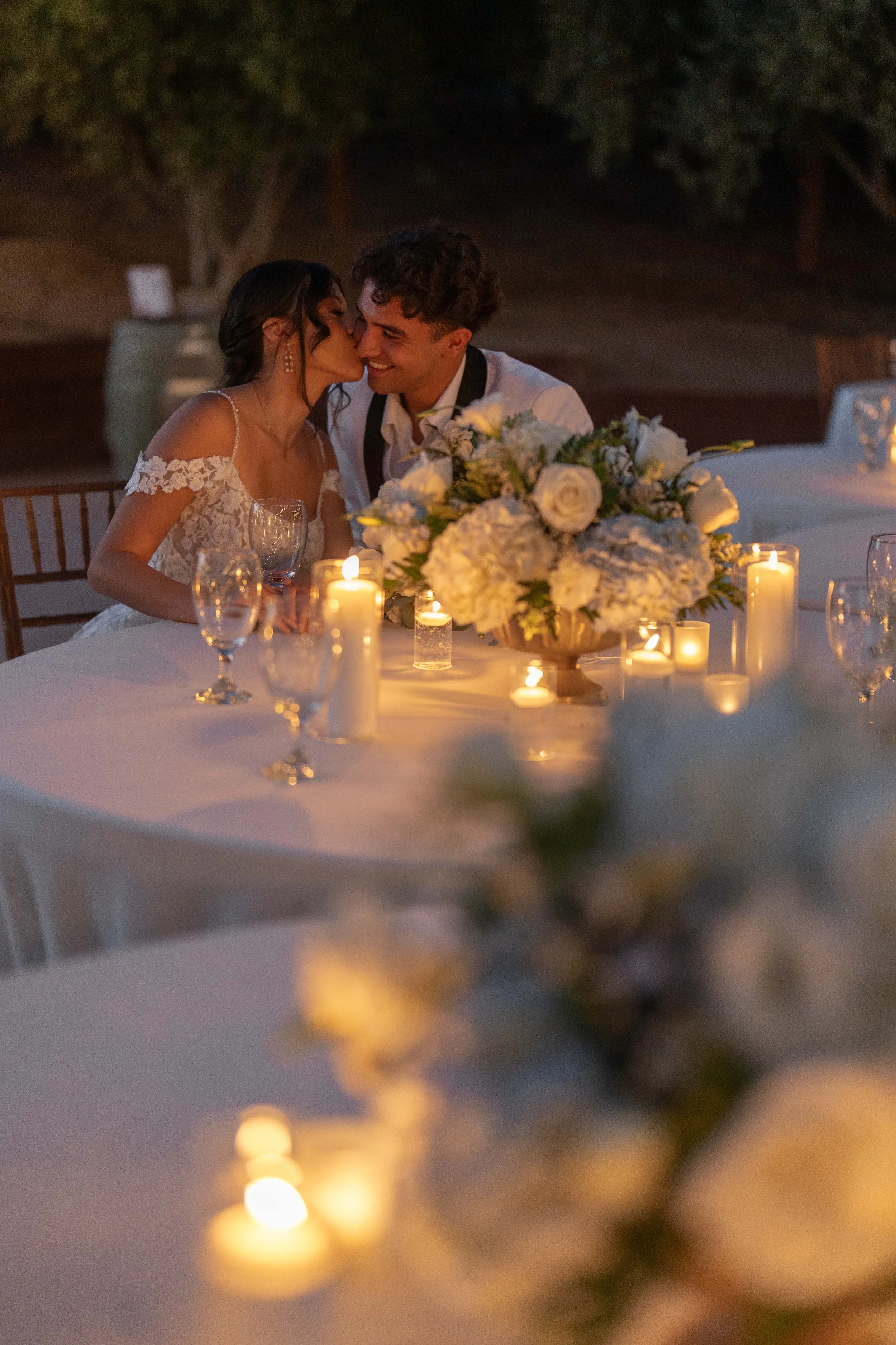 A couple sharing a romantic moment at a wedding dinner table decorated with candles and a large floral centerpiece in an outdoor setting at night.