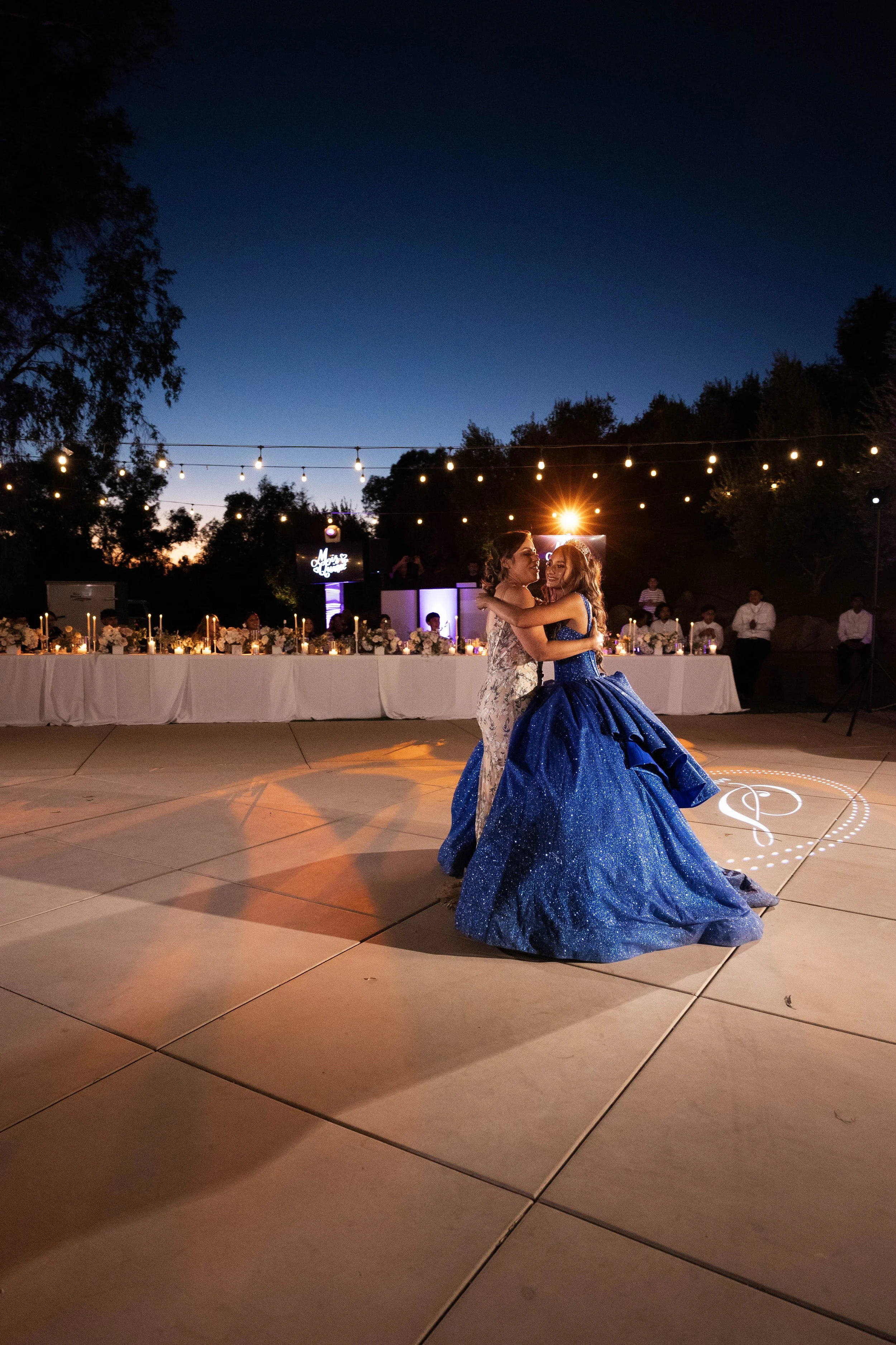 Two women in elegant dresses dancing together outdoors at night during a wedding reception, with a decorated long table, string lights, and a twilight sky in the background.