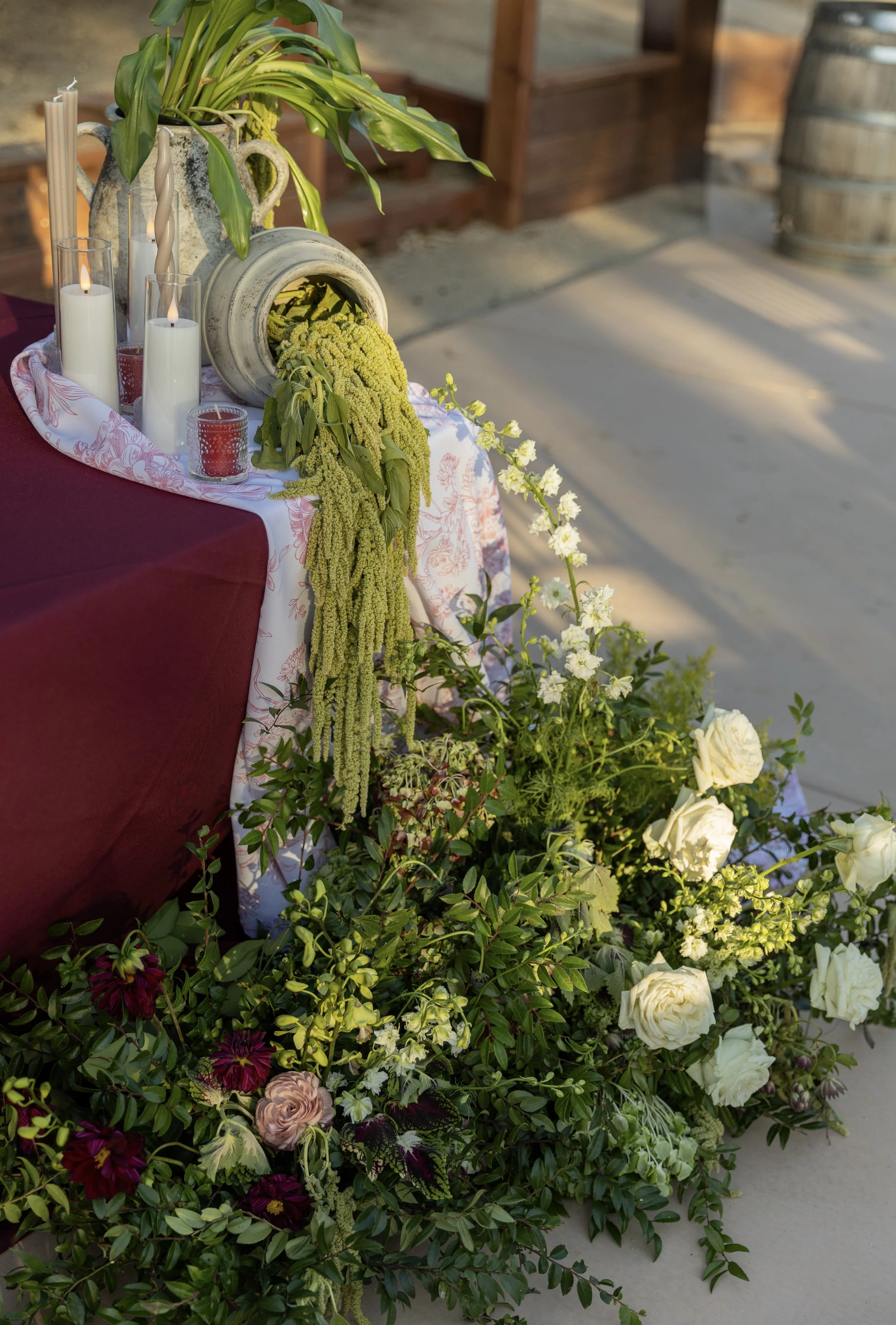 Decorative arrangement with candles, plants, and flowers on a table with a maroon tablecloth at an outdoor event.