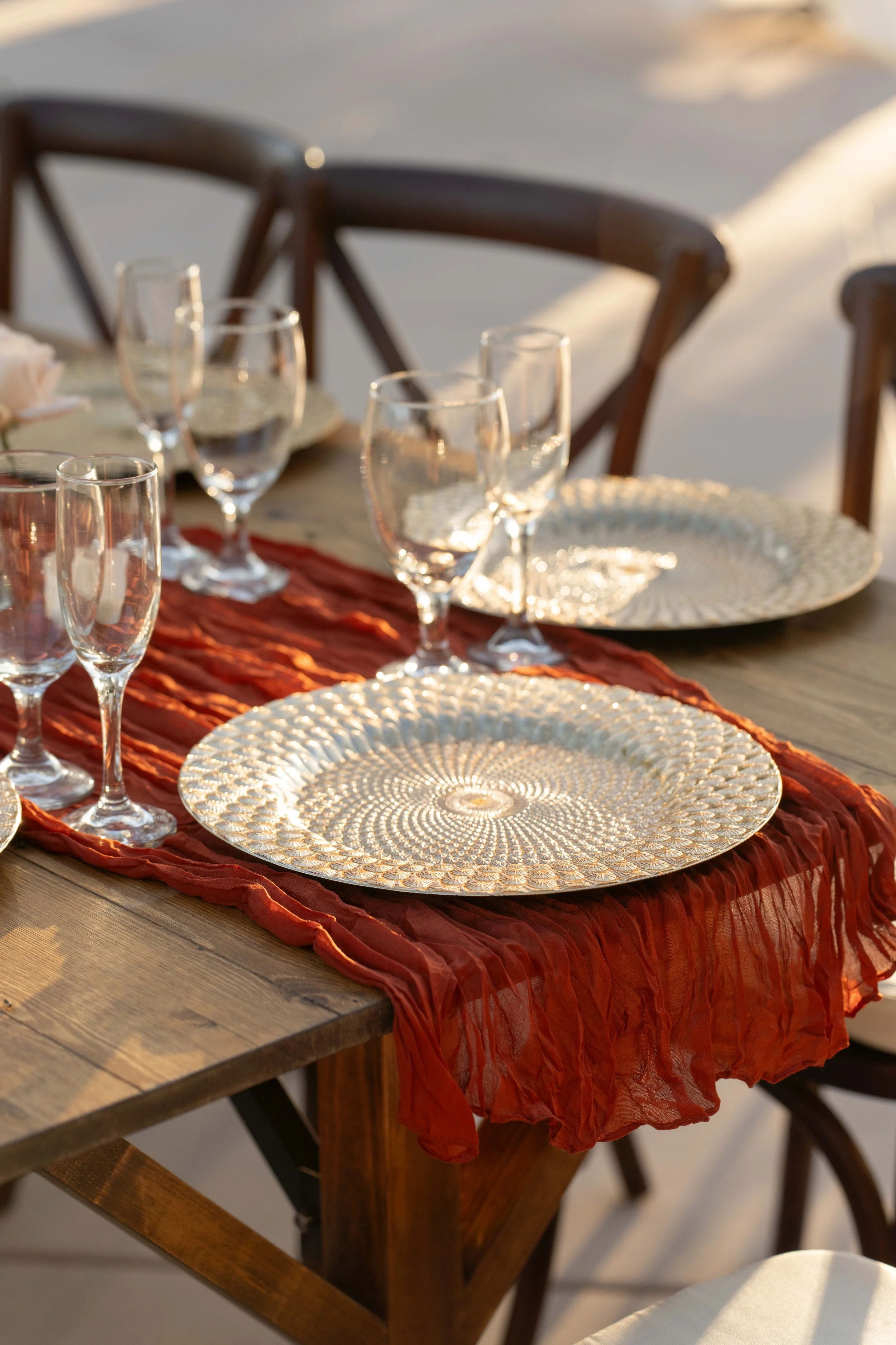 Elegant outdoor dining table set with gold patterned plates, clear wine glasses, and a red textured table runner at sunset.