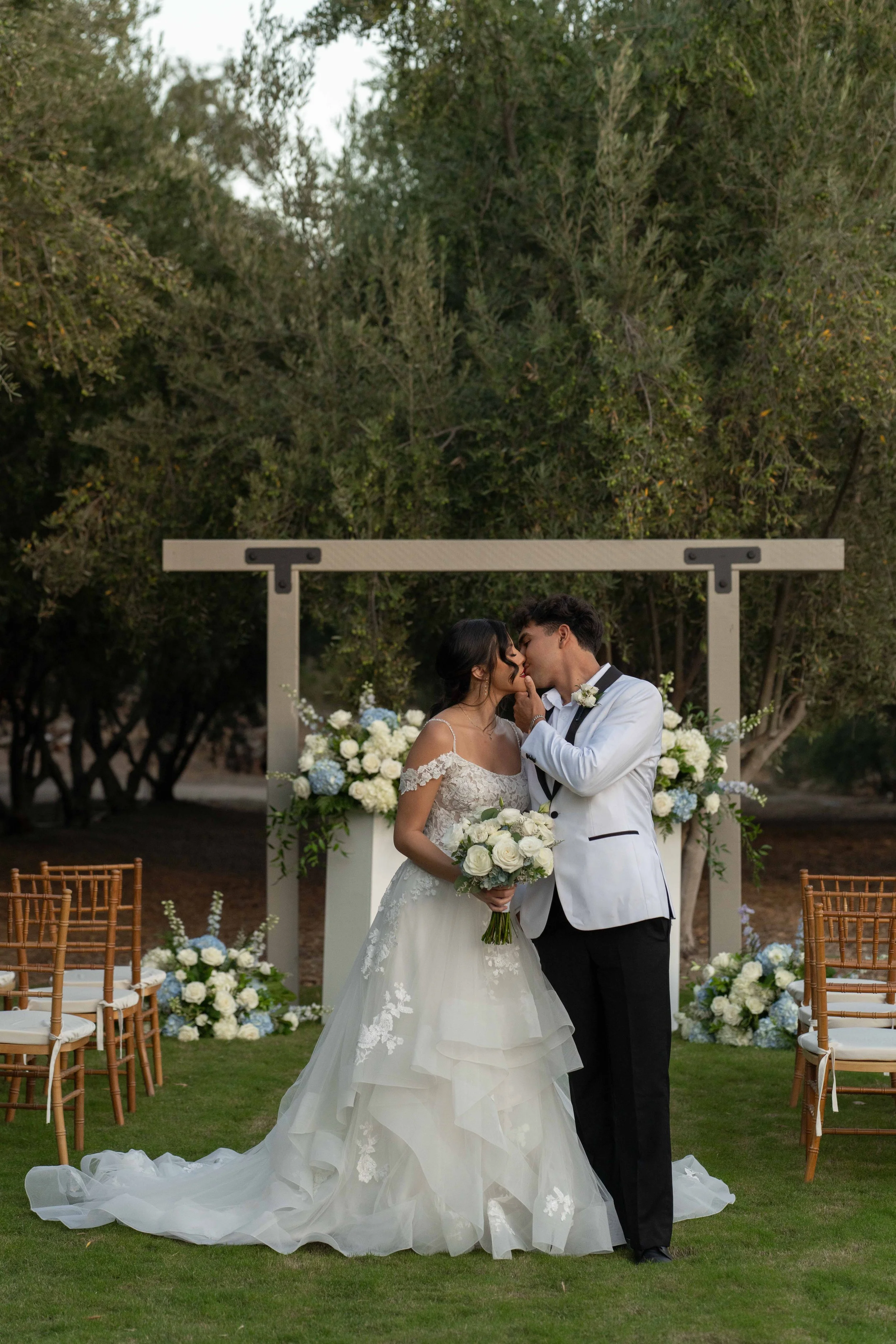 A bride and groom sharing a kiss at their outdoor wedding ceremony, with floral decorations and chairs around them.