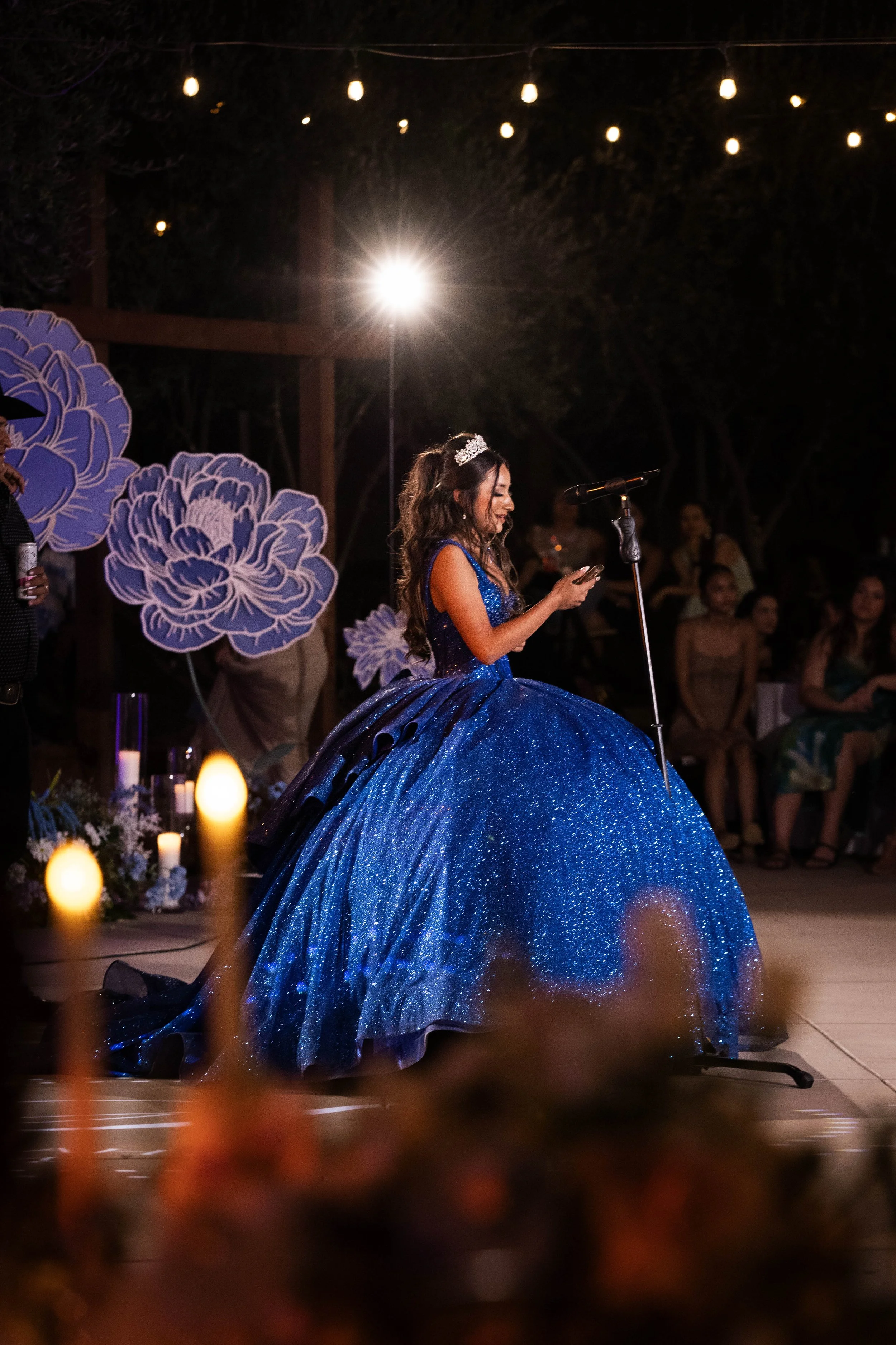 Young woman in a sparkly dark blue evening gown at an outdoor nighttime event, speaking into a microphone, with audience members in the background and decorative flowers and string lights overhead.