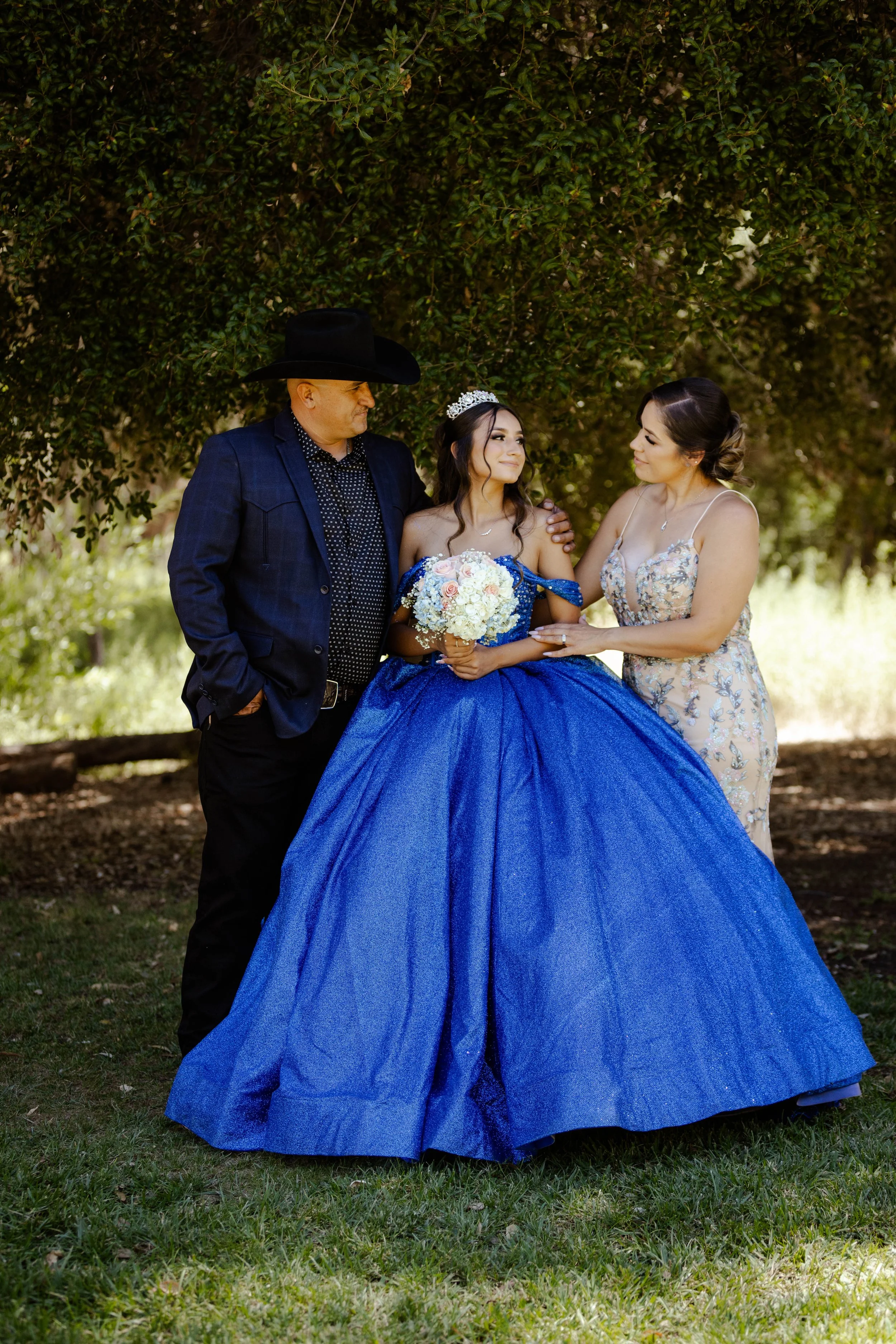 Young woman in a royal blue quinceañera dress holding a bouquet, standing with her parents under a tree, with the mother touching her arm and the father looking at her.