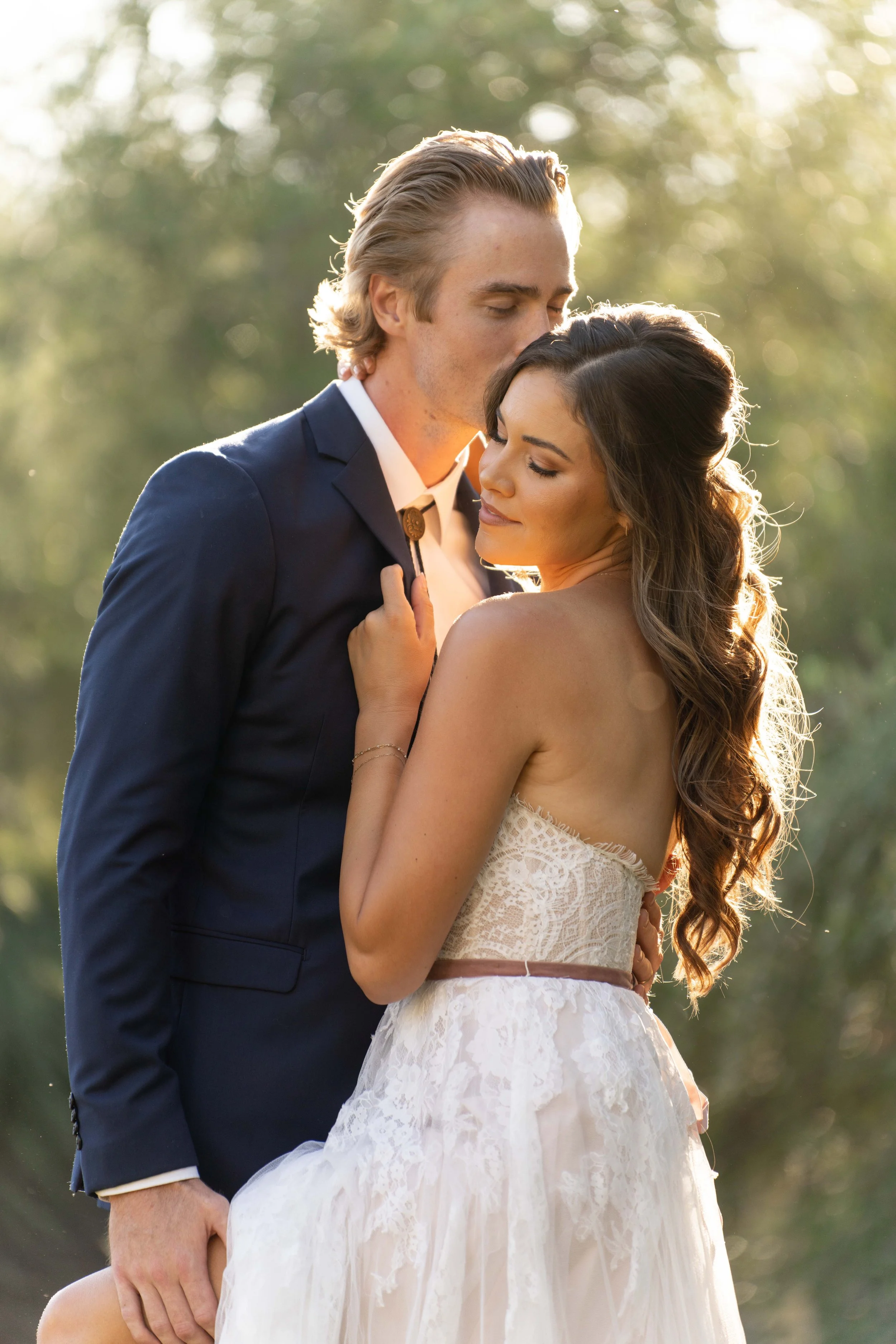 A loving couple embracing outdoors during sunset, with the man dressed in a navy suit and the woman in a lace wedding dress, both with eyes closed.