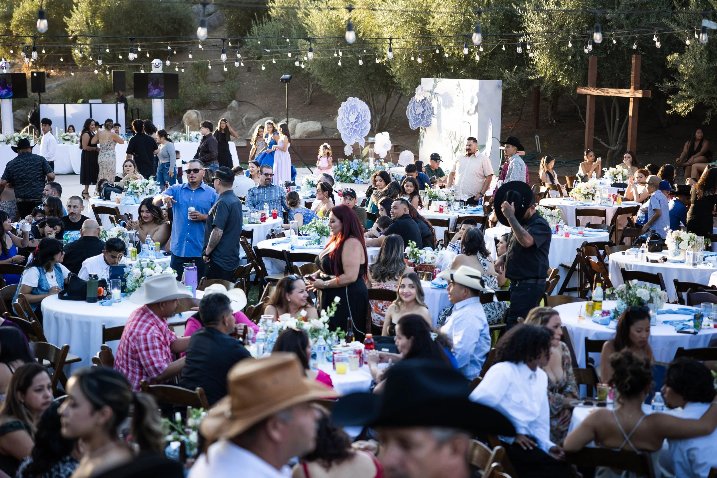 A lively outdoor celebration with numerous tables decorated with white and blue flowers, surrounded by guests socializing and enjoying drinks. String lights hang above, and a stage with decorations is in the background.