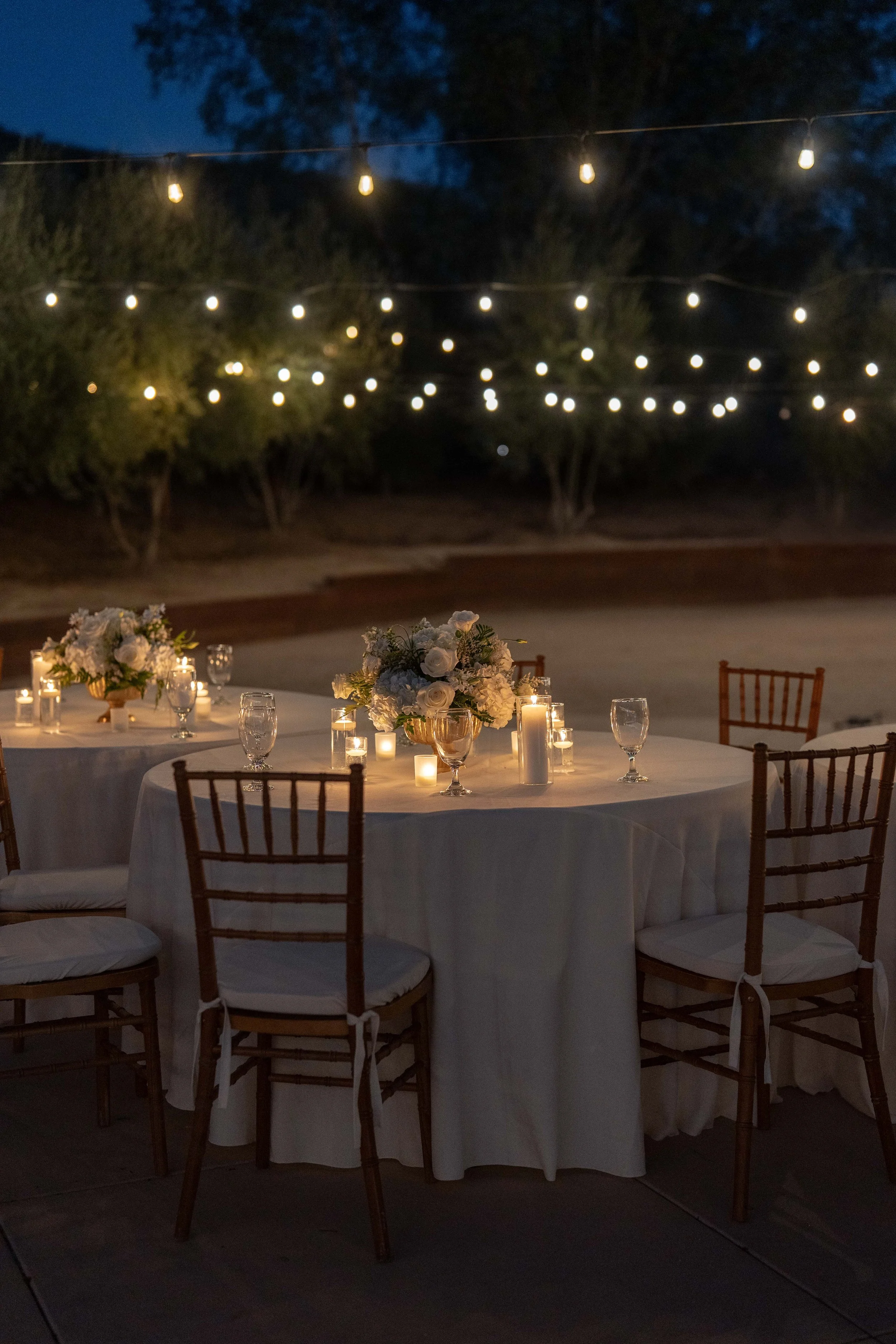 An outdoor evening event with a round table decorated with white floral arrangements and lit candles, under string lights in a natural setting.