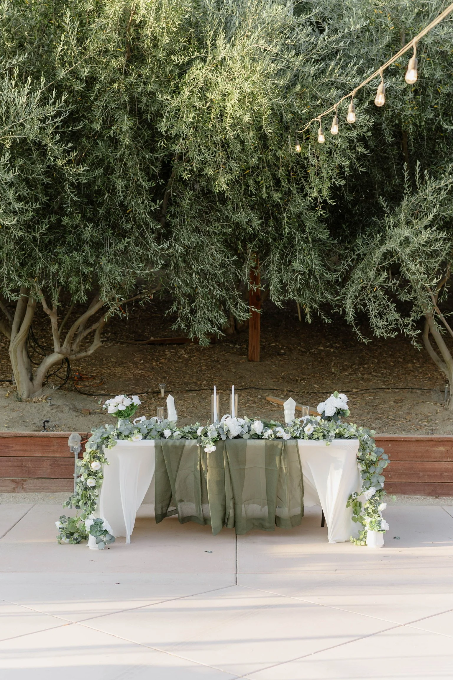 A decorated table set for an outdoor event, with white and green floral arrangements, candles, and green fabric accents, surrounded by trees and string lights.