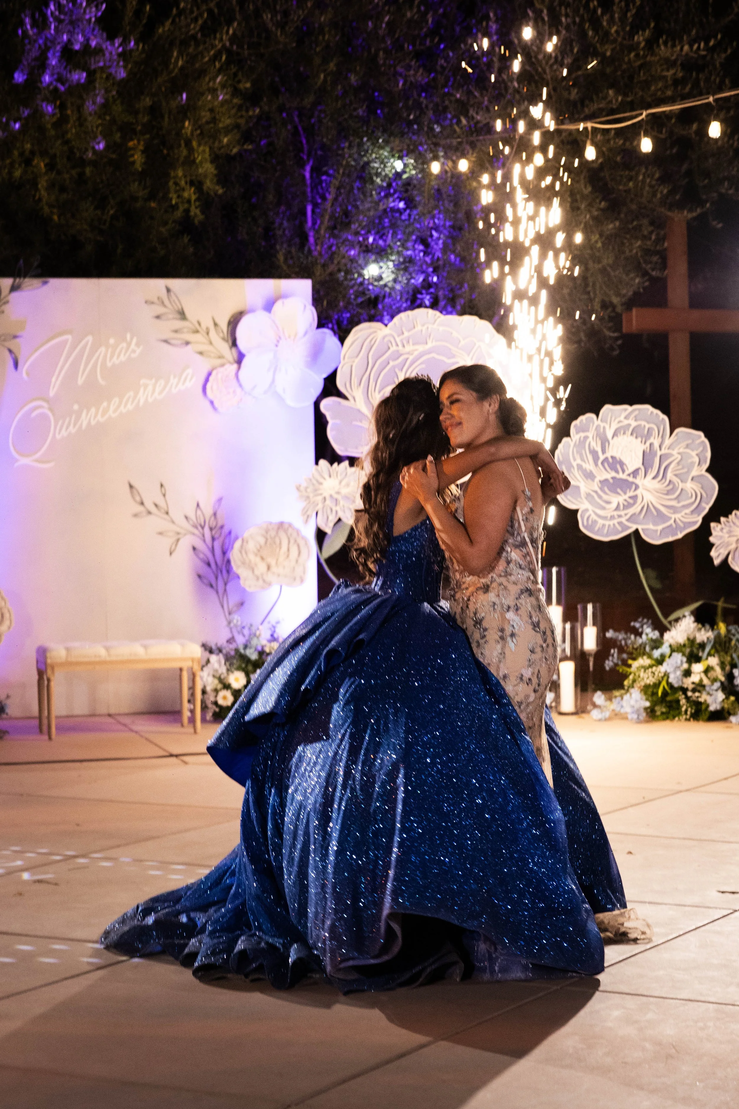 Two women dancing and embracing outdoors at a celebration, with a decorative backdrop and sparklers in the air.