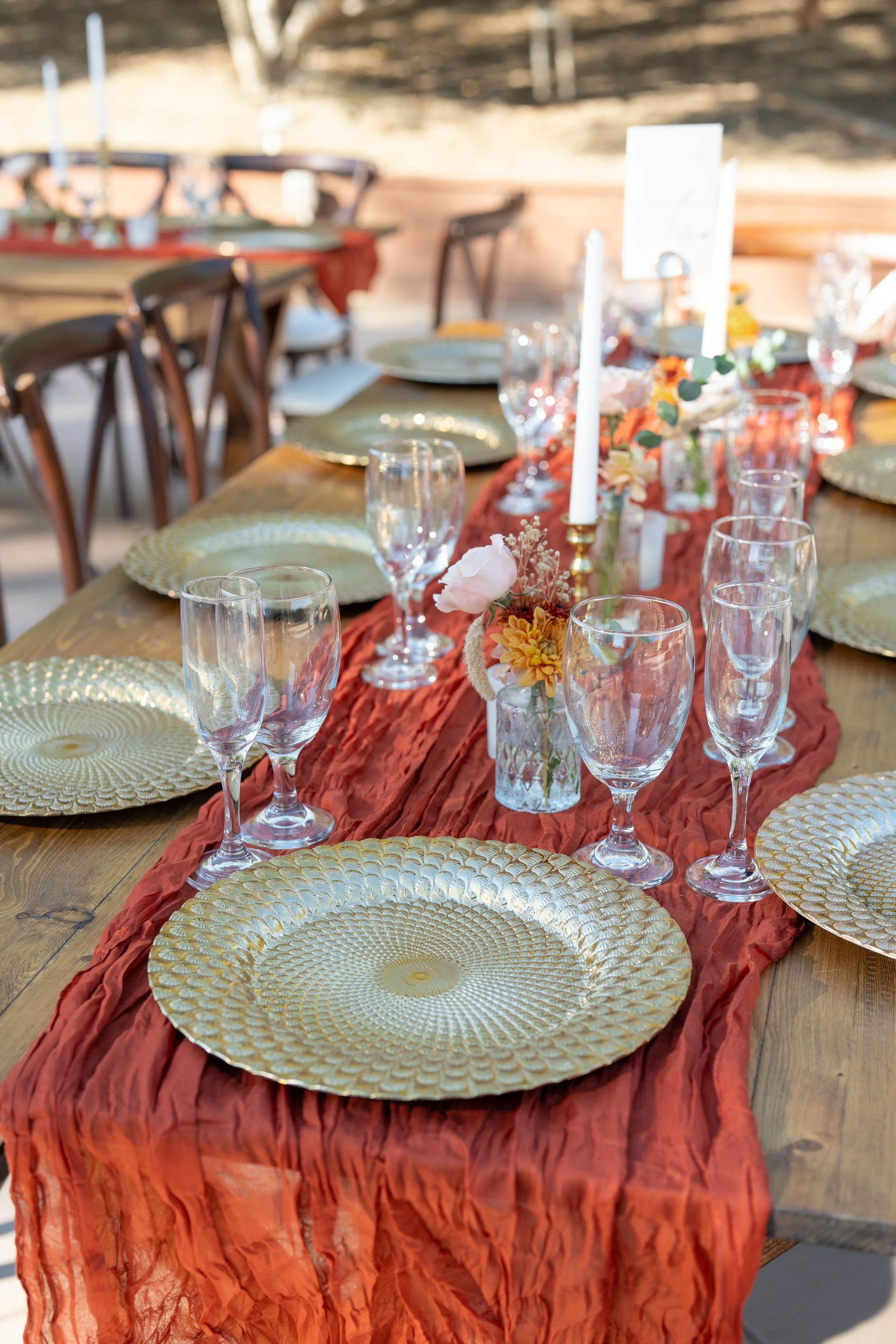 Set dining table with gold charger plates, clear glassware, white candles, and small floral arrangements on a rust-colored table runner.
