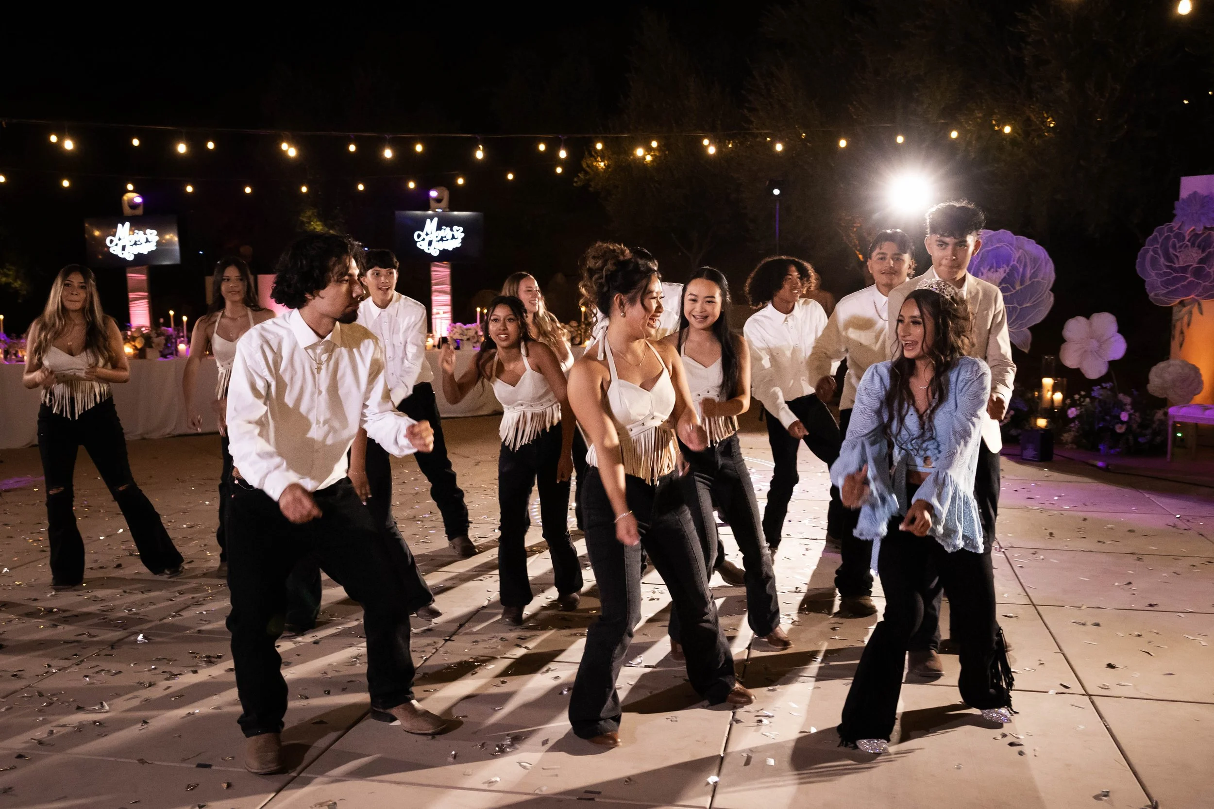 A group of young people dancing outdoors at night, illuminated by string lights and a bright spotlight, with decorated tables and large floral decorations in the background.