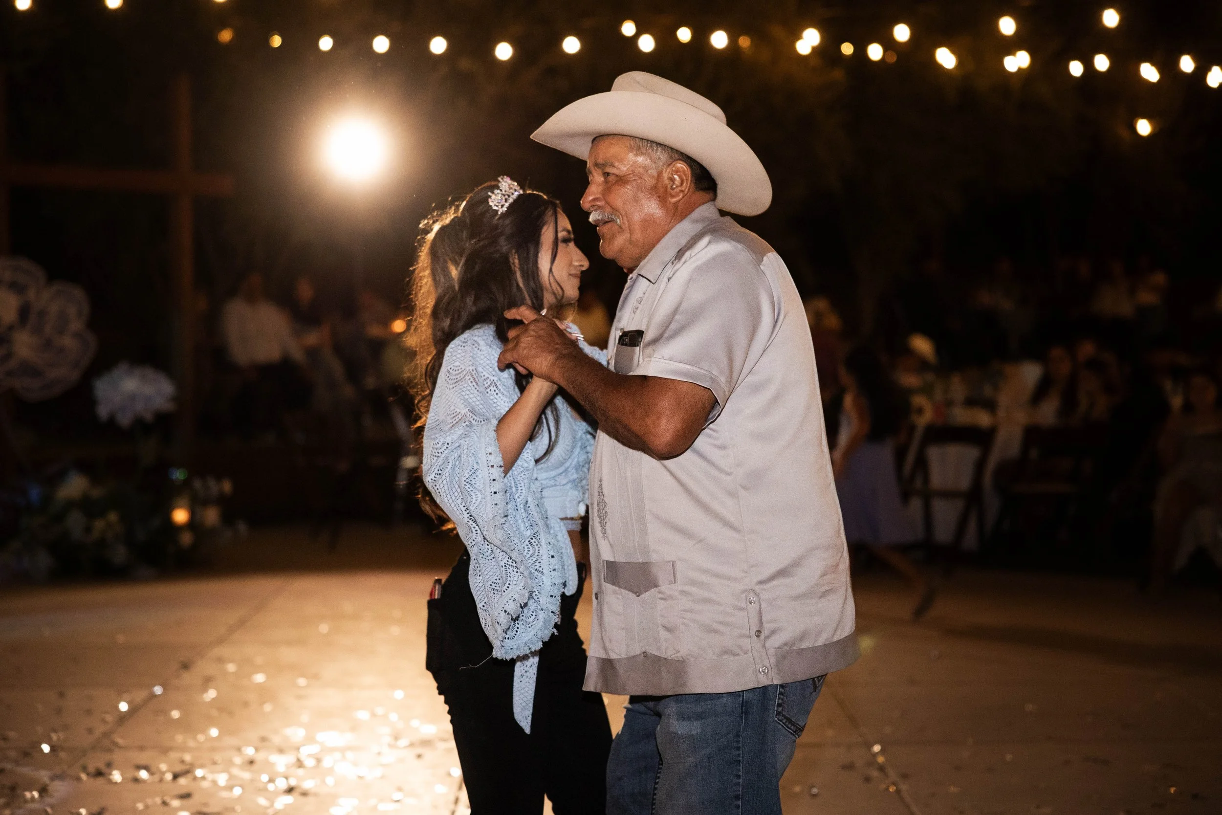 Older man and young girl dancing together at a party, with warm lights and blurred guests in the background.