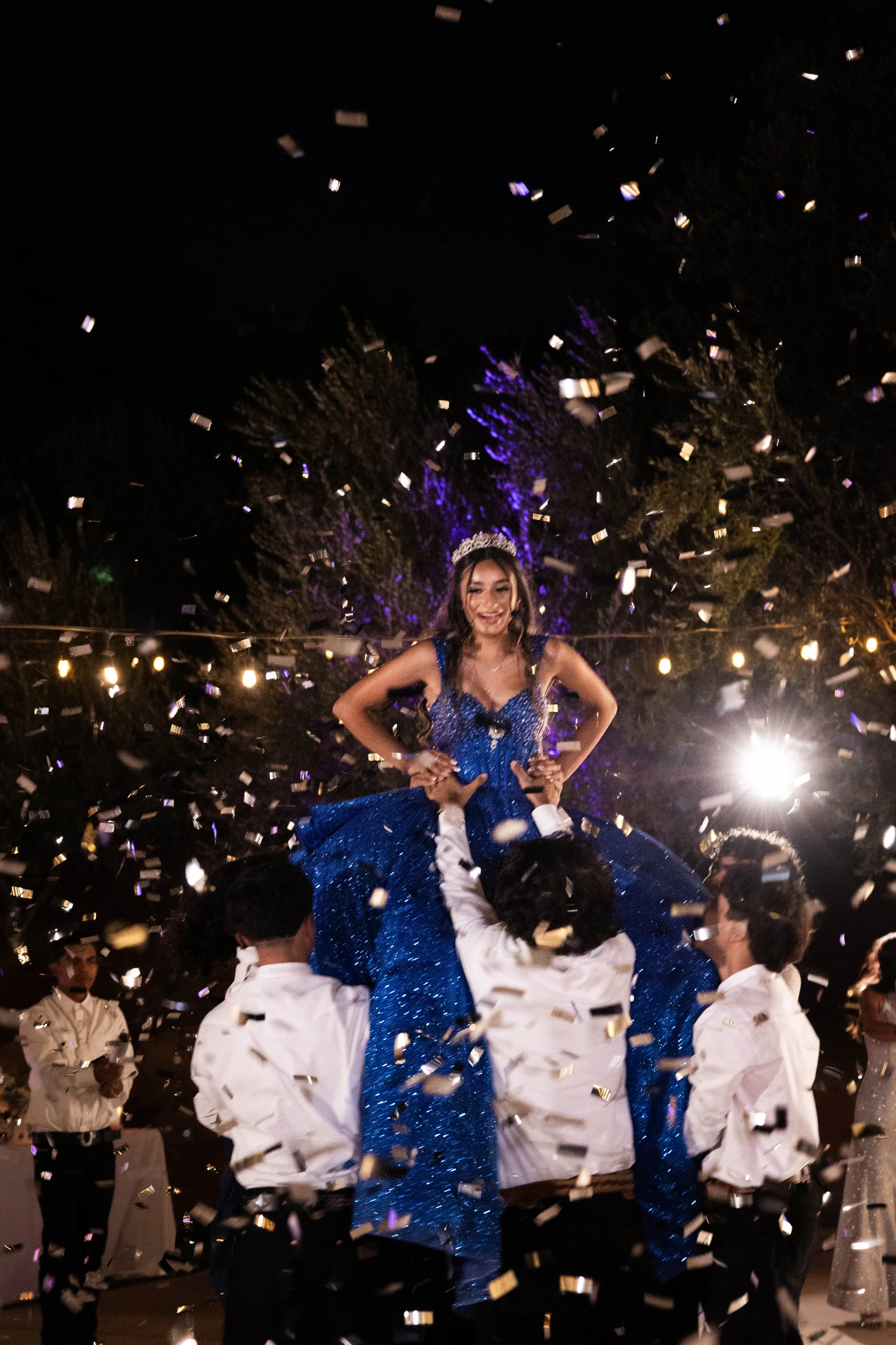 A girl in a blue dress being lifted by boys in white shirts during a celebration with confetti and string lights at night.