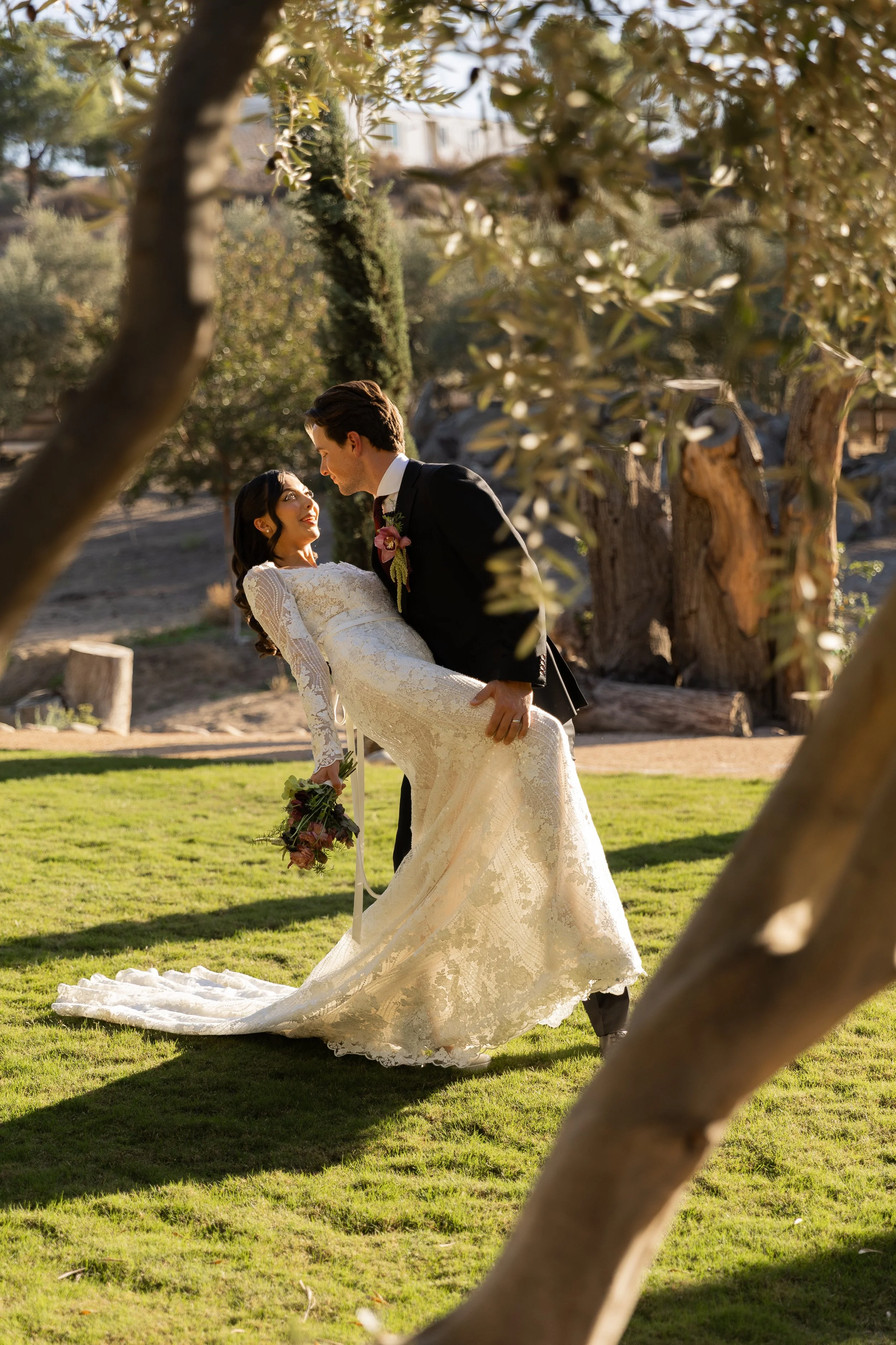 A groom is holding a bride in a white wedding dress in a park, with trees and greenery around, during what appears to be late afternoon sunlight.