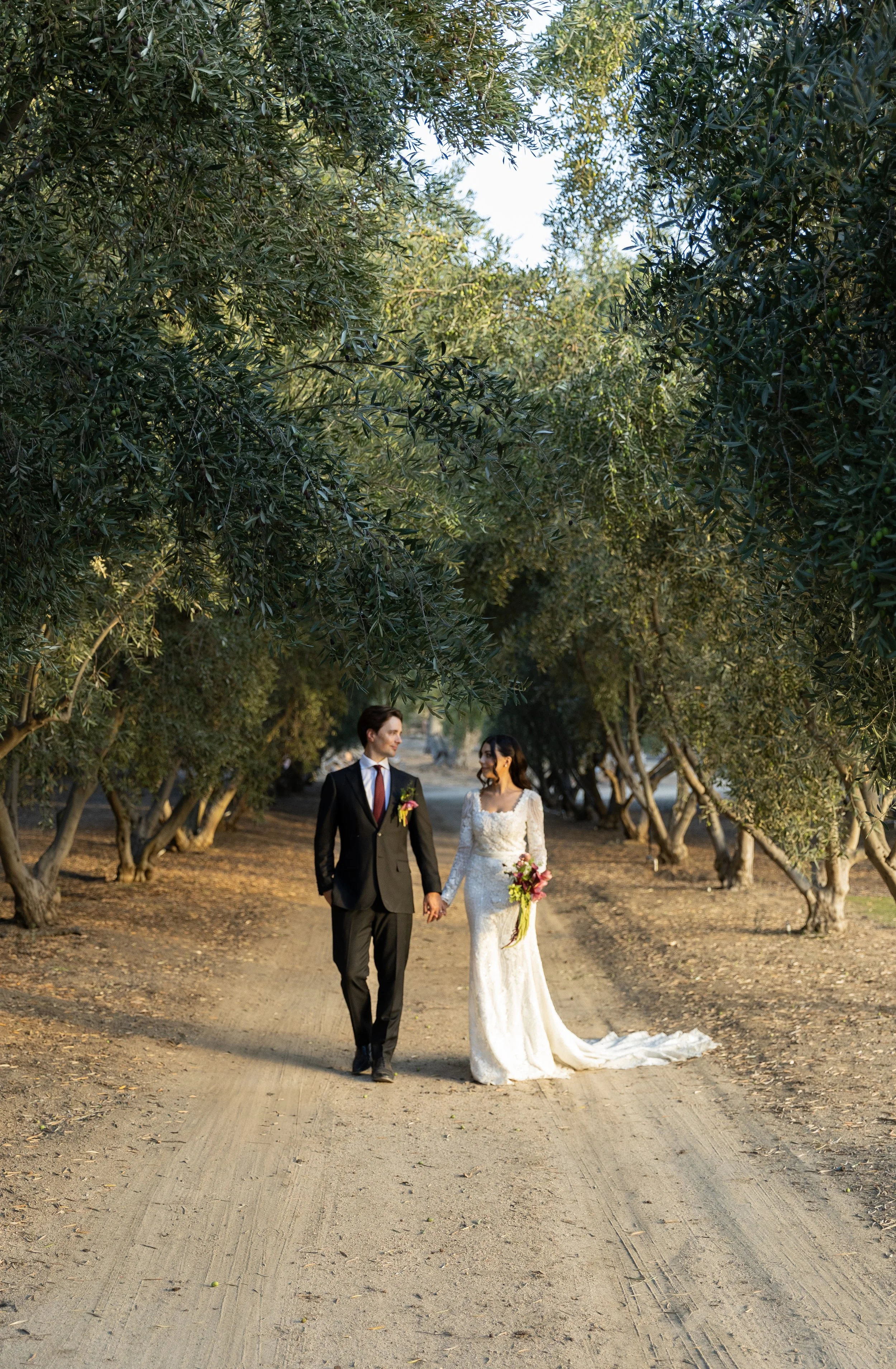 A bride and groom walking hand in hand on a dirt path lined with trees, during a wedding photoshoot, with lush green foliage overhead.