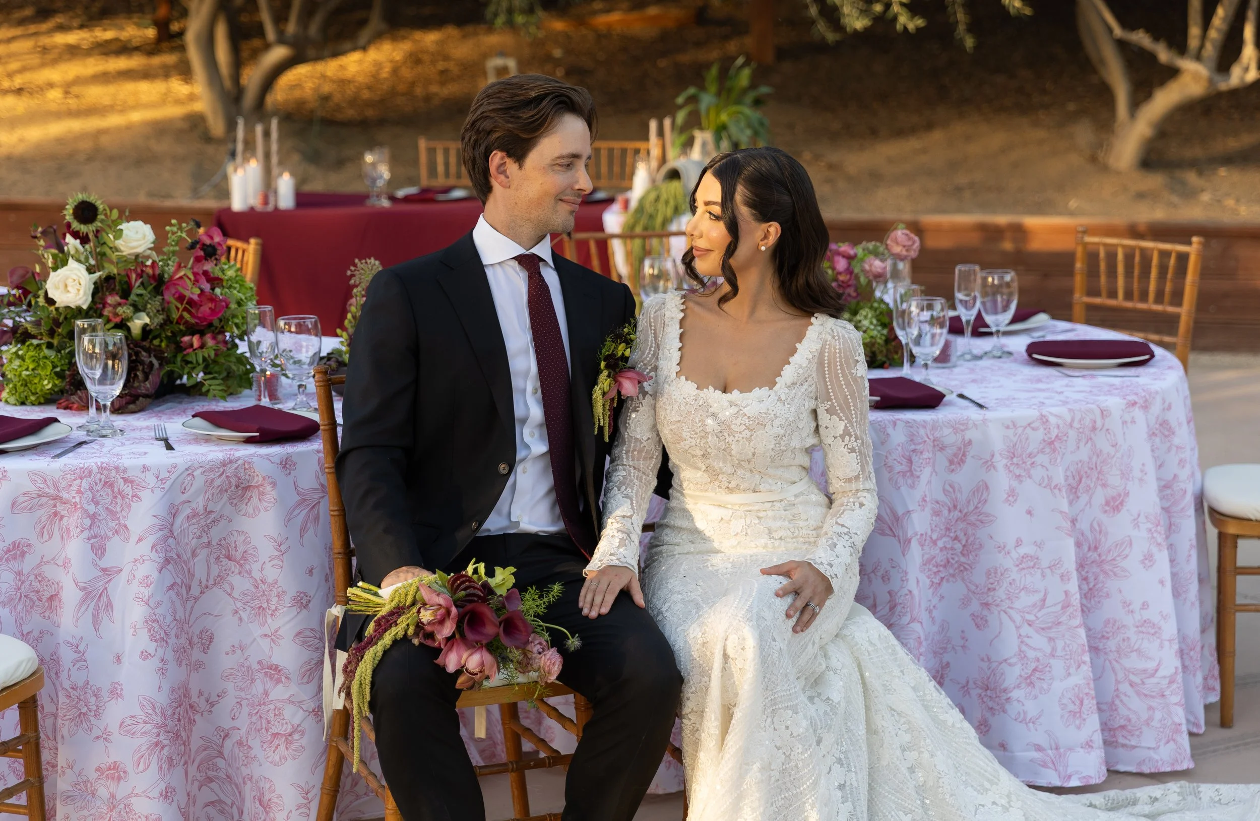 A bride and groom sitting close together at a wedding reception outdoors, surrounded by decorated tables with flowers and glassware.