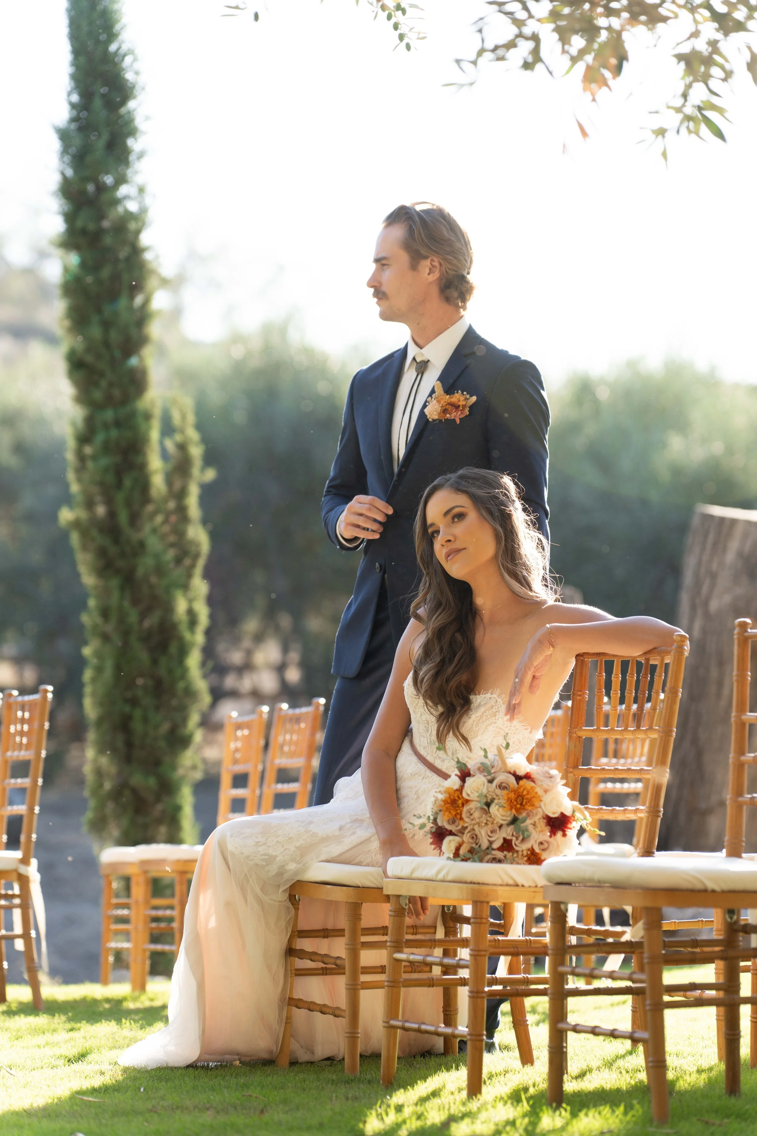 A bride and groom at an outdoor wedding ceremony. The bride, wearing a white strapless wedding dress, is seated on a wooden chair with a bouquet of flowers resting on her lap. The groom, wearing a dark suit with a boutonniere, stands beside her with 